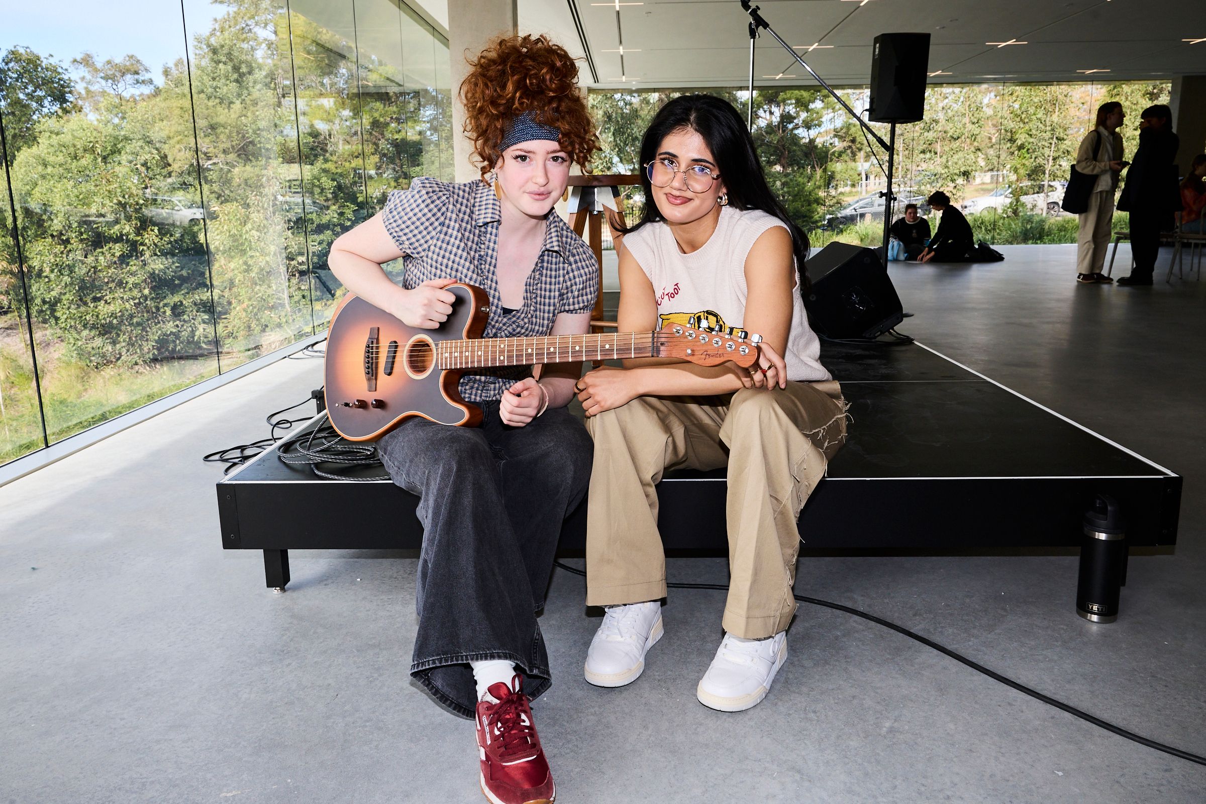 Guests and musicians Maya Riv (left) and Ayesha Madon (right), posed sitting on the side of a stage with a guitar.