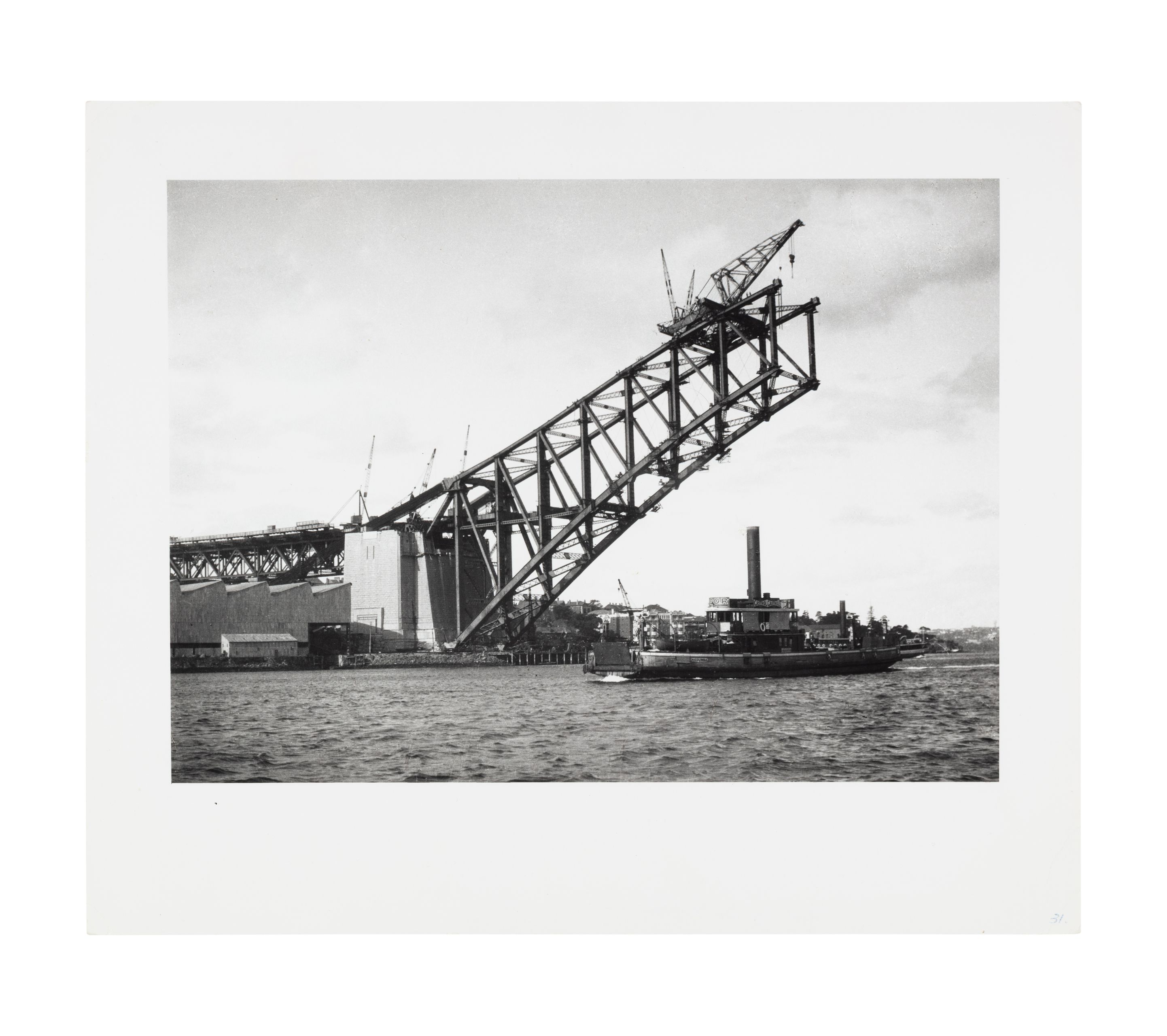 Image of one side of the harbour bridge arch and pylon under construction. Cranes are visible at the top of the arch and the road level above the pylon. A ferry is visible below the structure on the harbour.