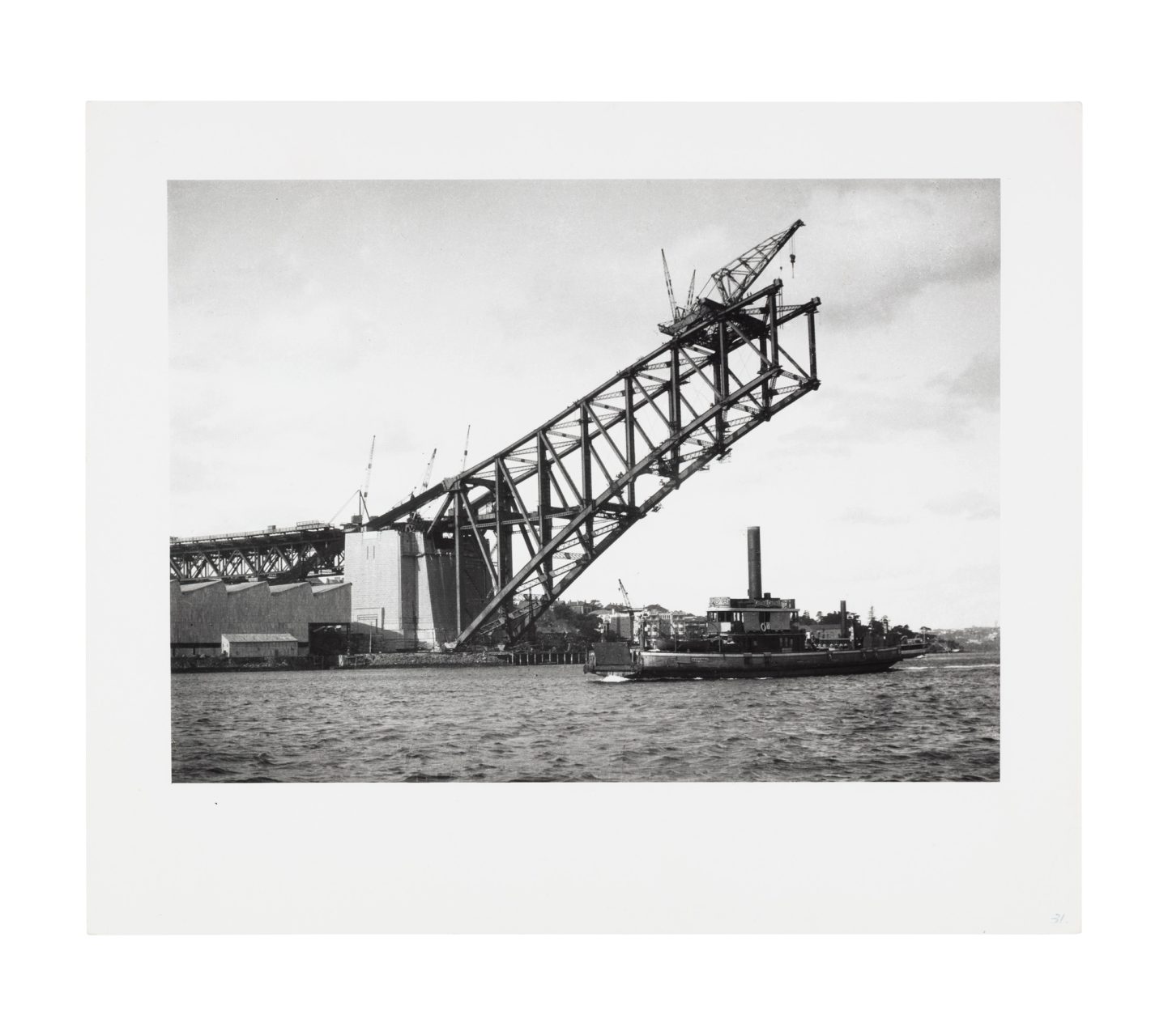 Image of one side of the harbour bridge arch and pylon under construction. Cranes are visible at the top of the arch and the road level above the pylon. A ferry is visible below the structure on the harbour.