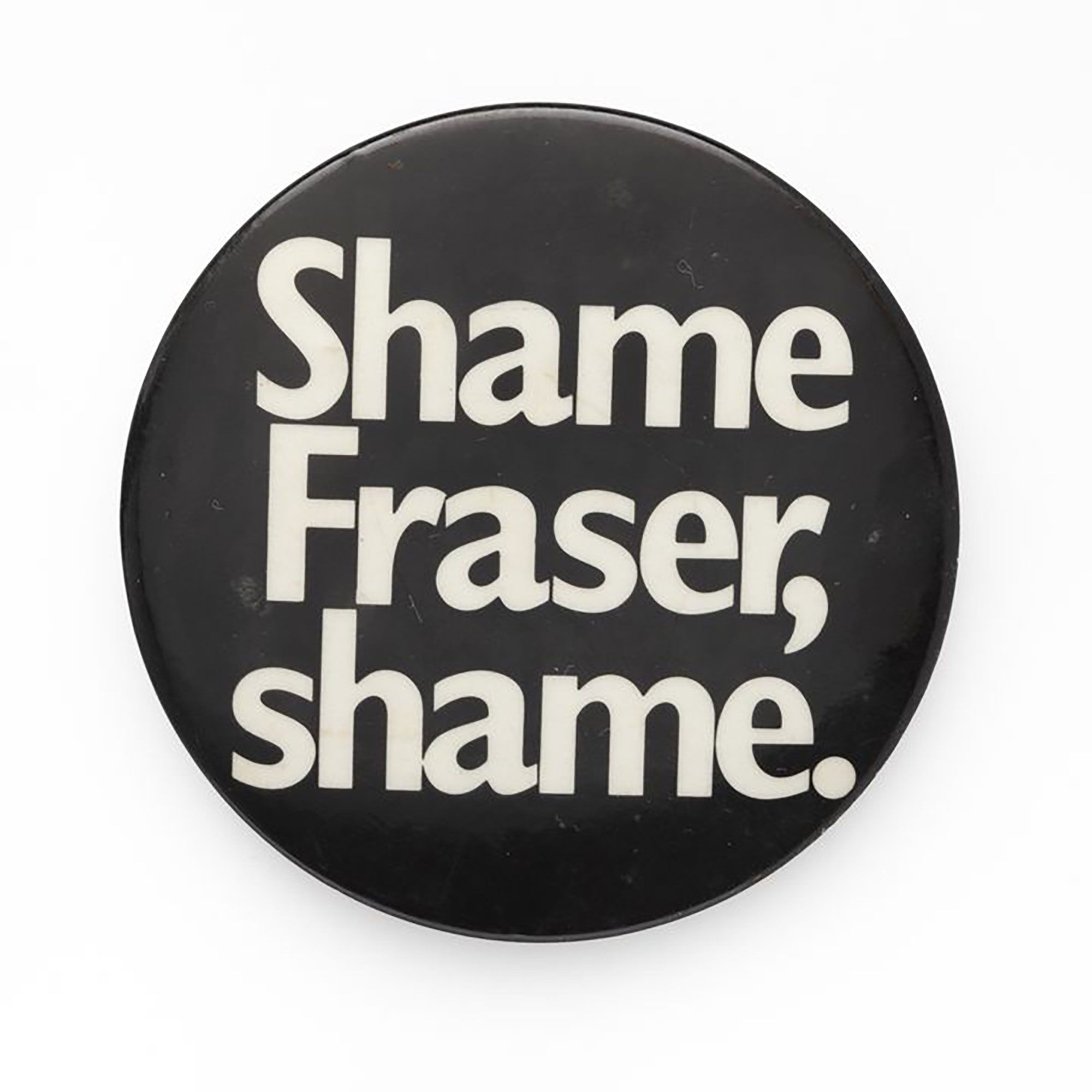 Circular metal badge, front with black ground and white lettering 'Shame Fraser, Shame.'. Verso white paint and pin.
