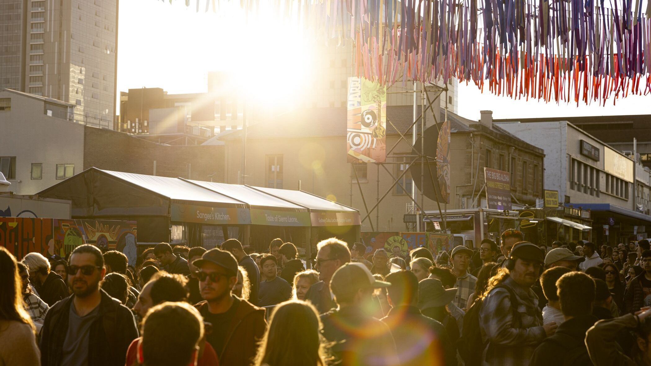 A bustling outdoor market scene filled with a crowd of people, illuminated by the low-lying sun, and with colourful streamer decorations overhead.