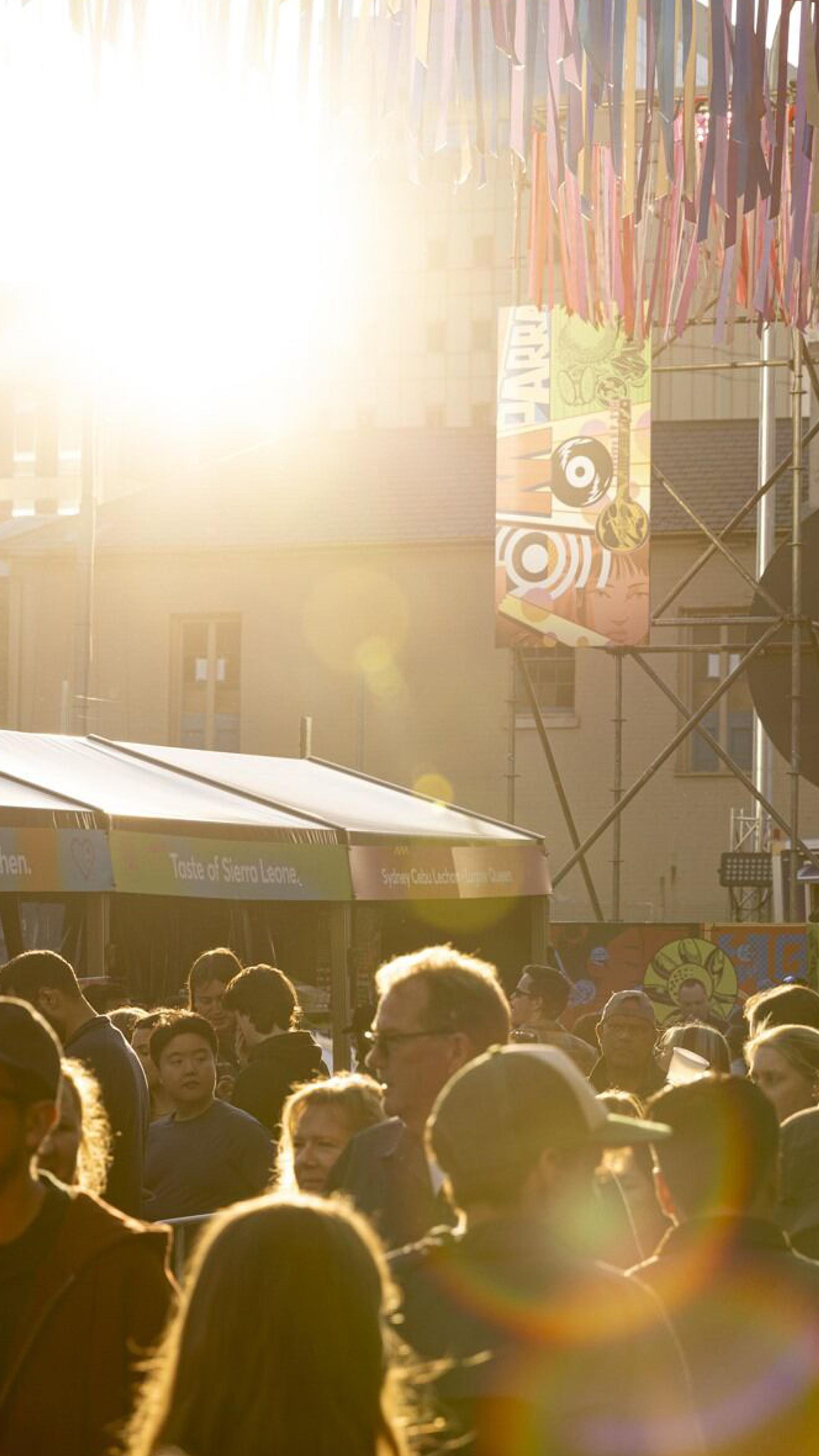 A bustling outdoor market scene filled with a crowd of people, illuminated by the low-lying sun, and with colourful streamer decorations overhead.