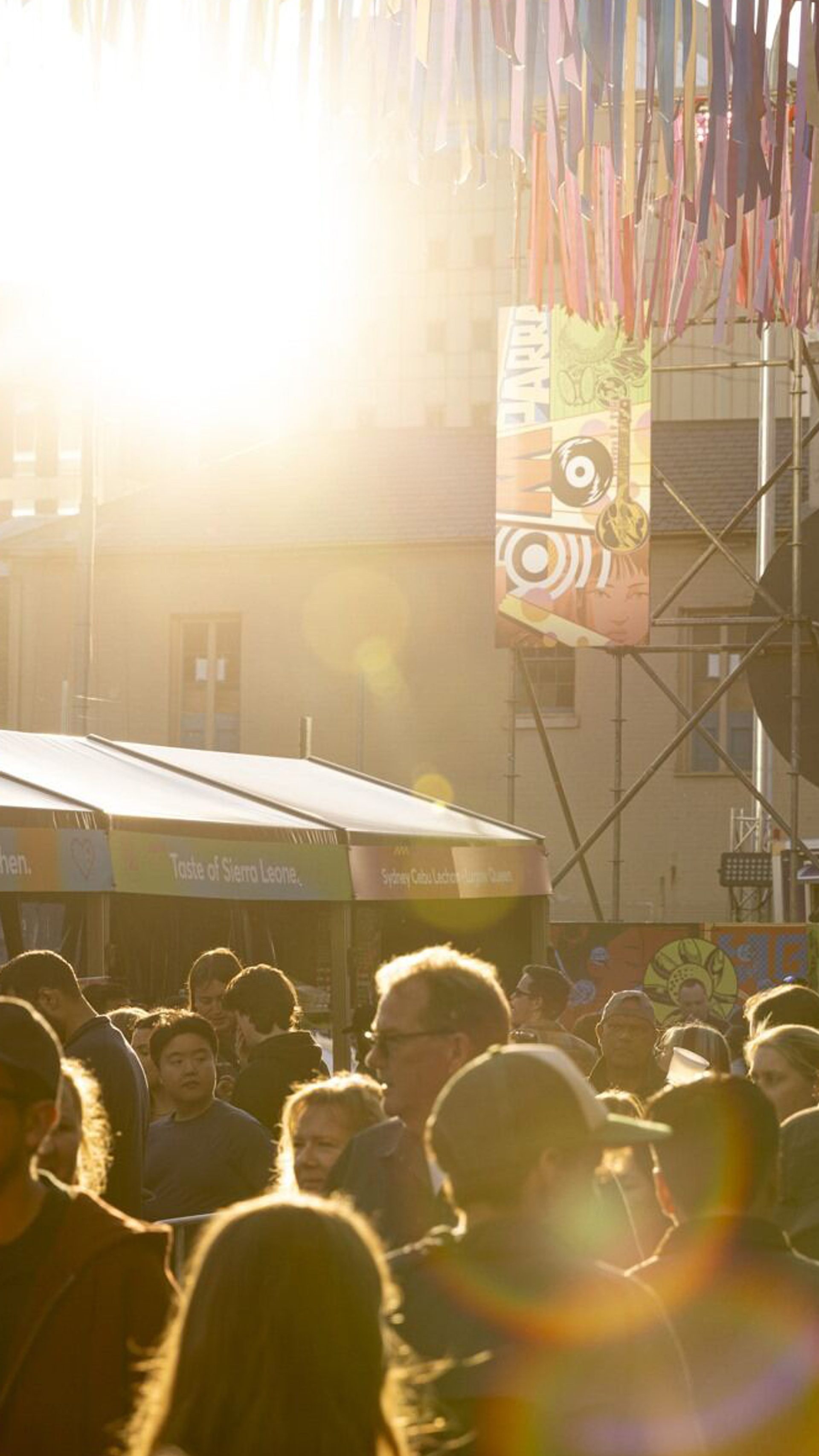 A bustling outdoor market scene filled with a crowd of people, illuminated by the low-lying sun, and with colourful streamer decorations overhead.