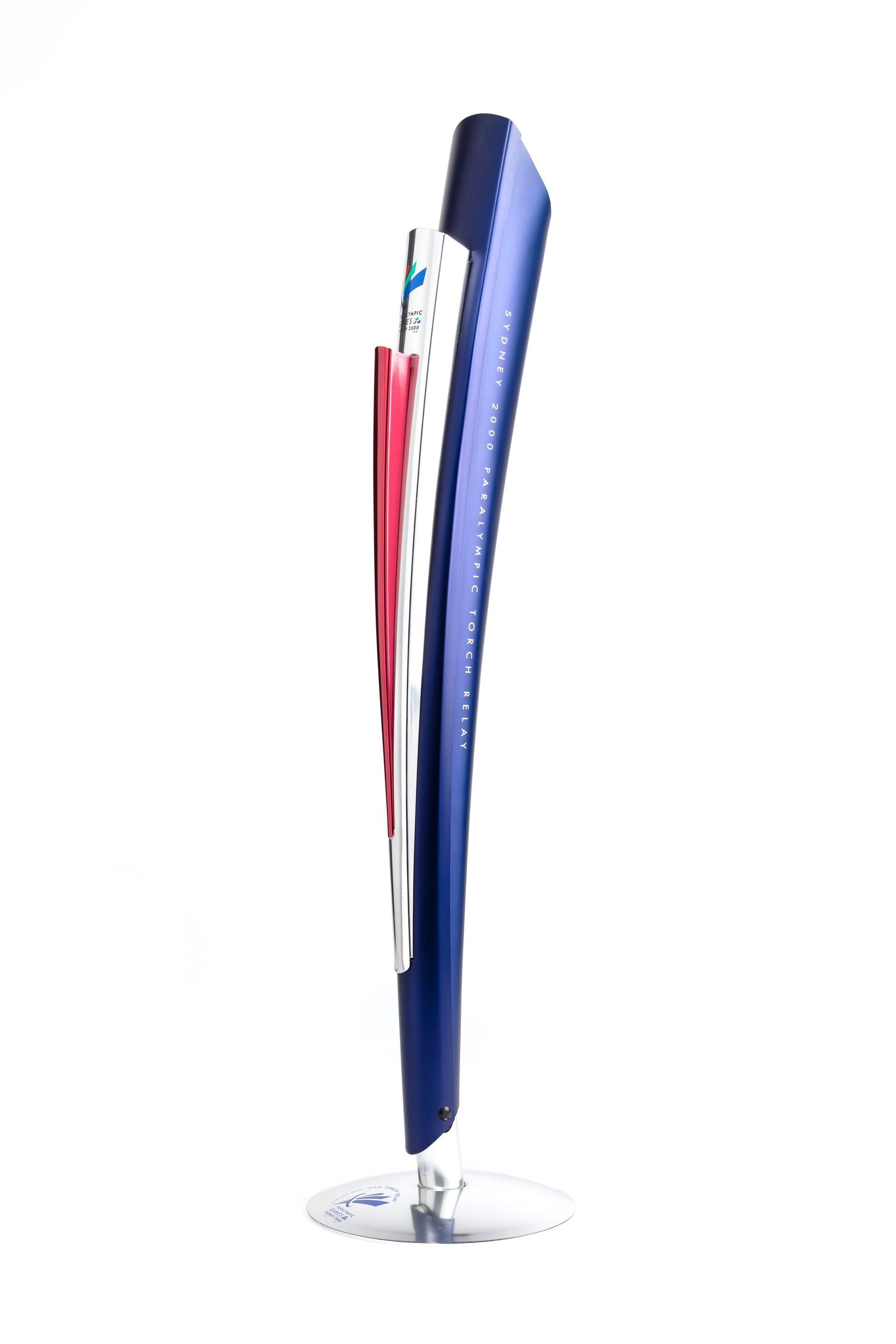 Paralympics torch made of three tapered cylinders of shiny metal (red, silver and blue) on a silver stand. Text on the blue piece reads: 'Sydney 2000 Paralympic Torch Relay'.