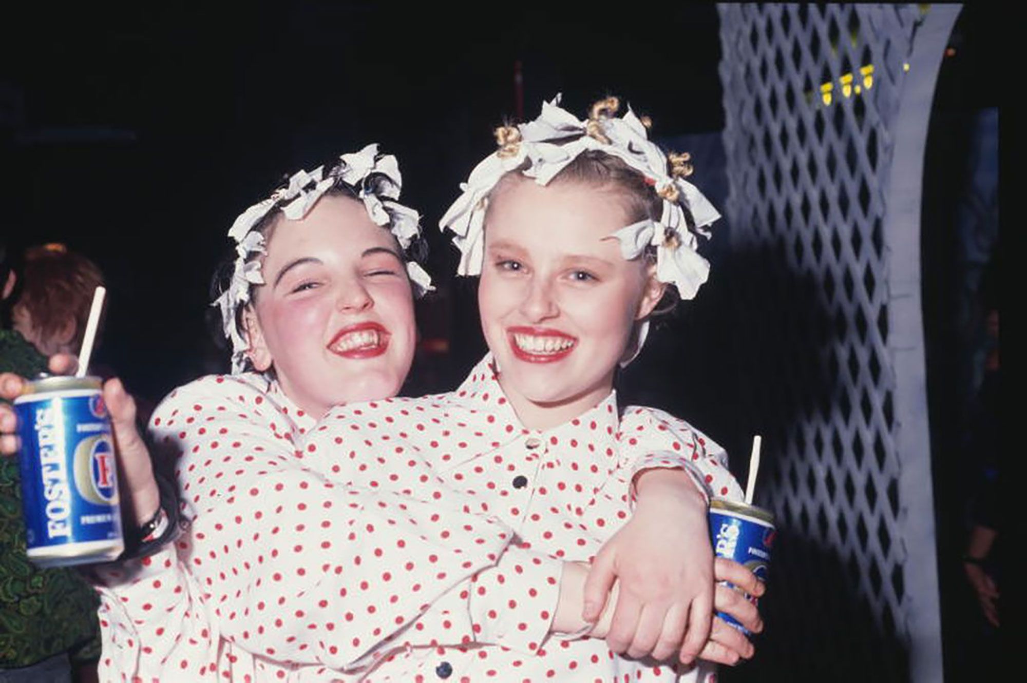 Two young women wearing matching polka dot shirts (white with red dots) smile at the camera. They are each holding a can of Fosters beer, with a straw. Both women are wearing red lipstick and have curling rags in their hair.