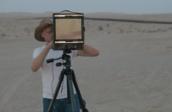 A man in dark trousers, white T-shirt and hat looks through a screen on a tripod as he stands in a desert-like environment.