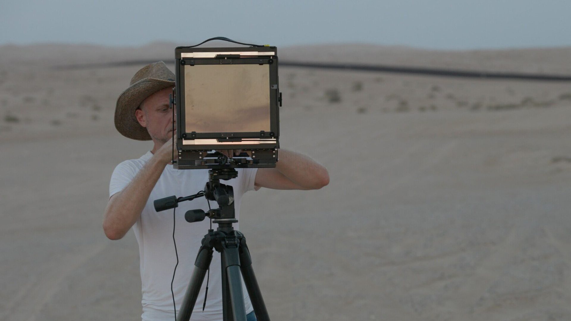 A man in dark trousers, white T-shirt and hat looks through a screen on a tripod as he stands in a desert-like environment.
