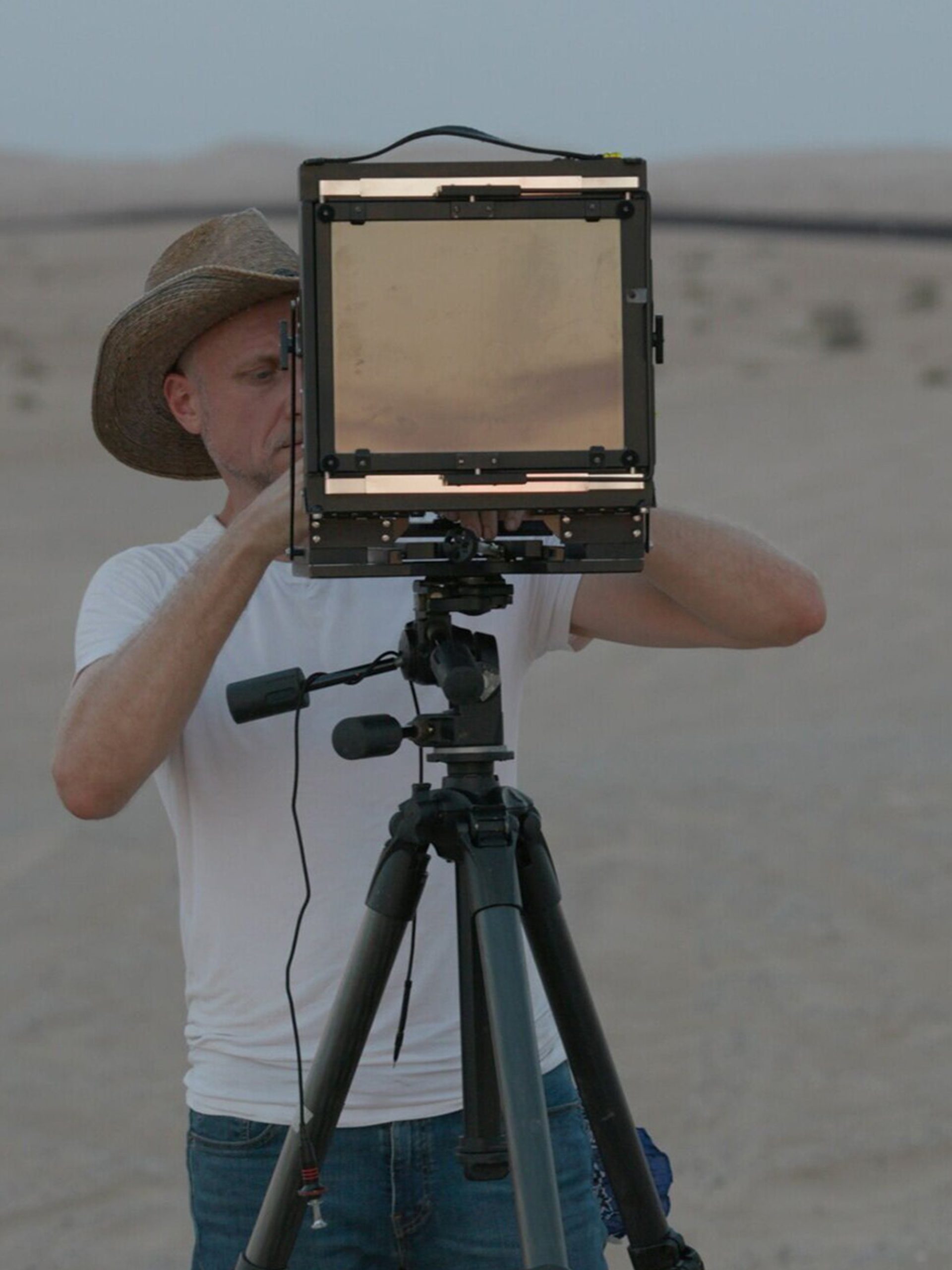A man in dark trousers, white T-shirt and hat looks through a screen on a tripod as he stands in a desert-like environment.