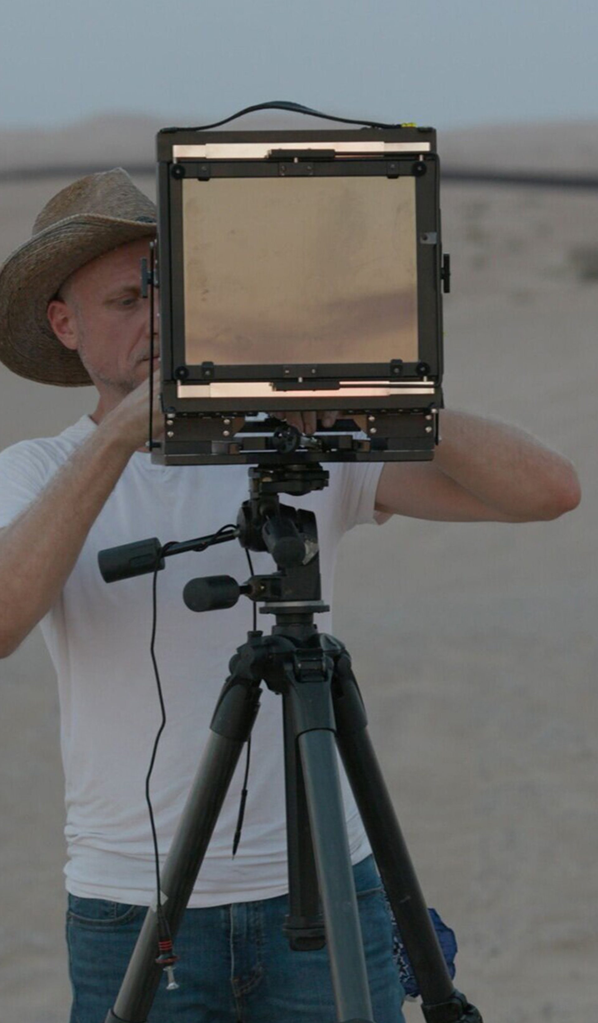 A man in dark trousers, white T-shirt and hat looks through a screen on a tripod as he stands in a desert-like environment.