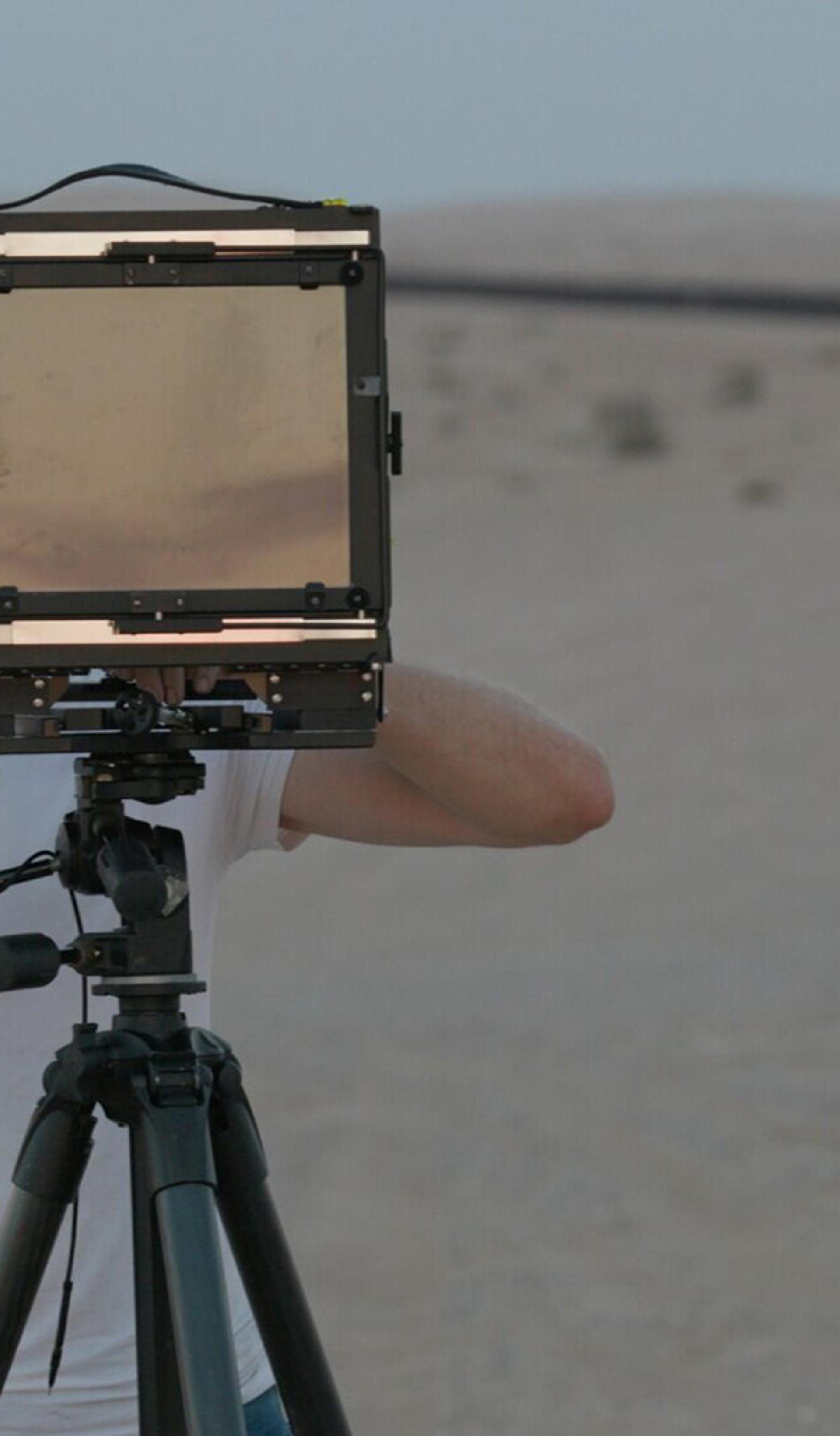 A man in dark trousers, white T-shirt and hat looks through a screen on a tripod as he stands in a desert-like environment.