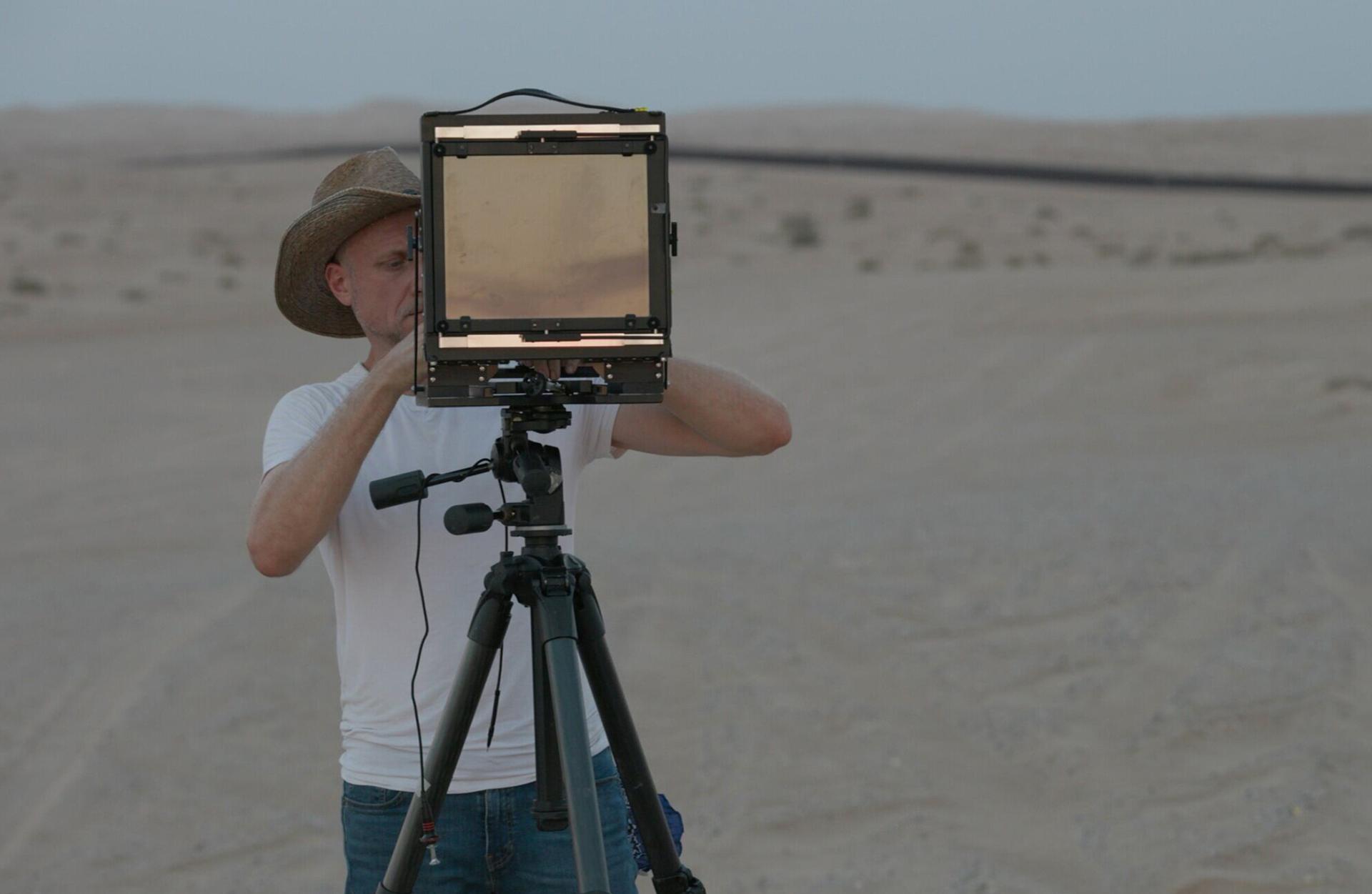 A man in dark trousers, white T-shirt and hat looks through a screen on a tripod as he stands in a desert-like environment.