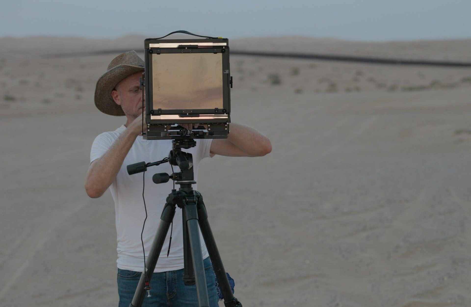 A man in dark trousers, white T-shirt and hat looks through a screen on a tripod as he stands in a desert-like environment.