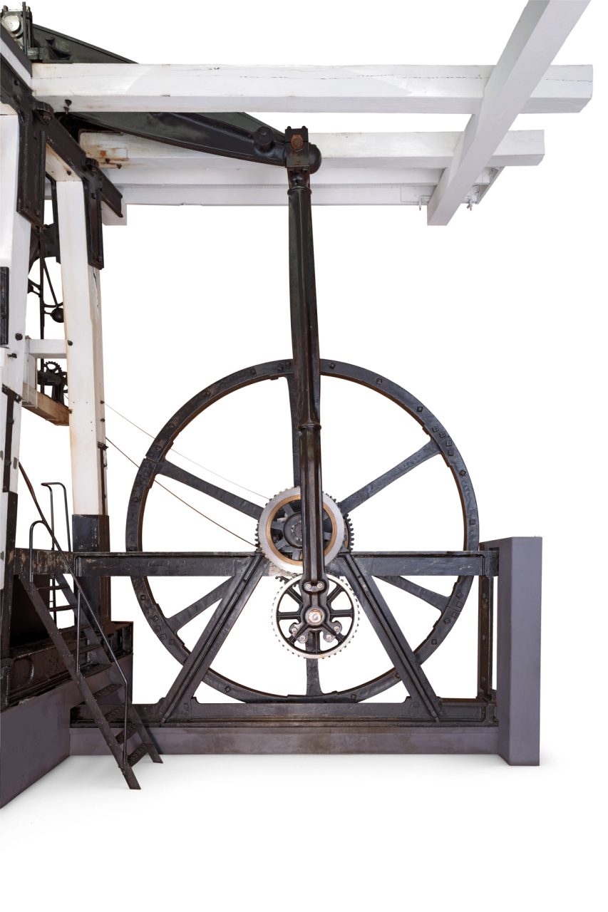 Photograph of one side of a steam engine. The vertical cylinder is connected to a separate condensor to which spent steam is exhausted and cooled to form water, creating a partial vacuum. The piston, which is pushed up and then down by steam admitted to the cylinder below and then above it, is attached to the overhead beam. The other end of the beam transfers motion via a connecting rod to the 'planet' gear, which turns the horizontal drive shaft via the 'sun' gear. A large flywheel, which maintains momentum between strokes, is attached to the horizontal drive shaft, which also carries the smaller toothed drive wheel and the governor pulley.