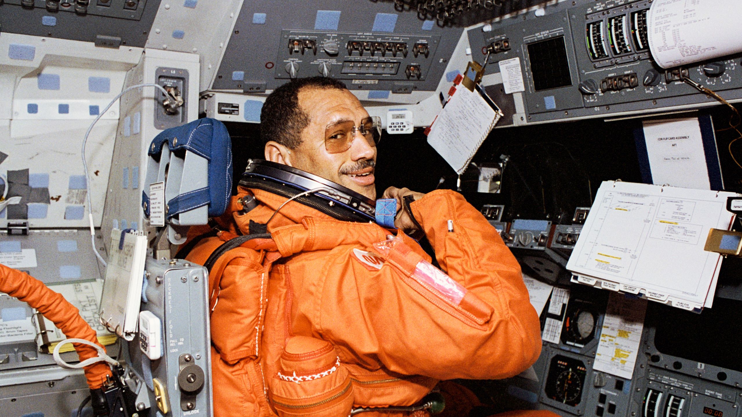 An astronaut commander (Charles F Bolden Jr) in a bright orange spacesuit sitting on the left station on the forward flight deck of the space shuttle Discovery.