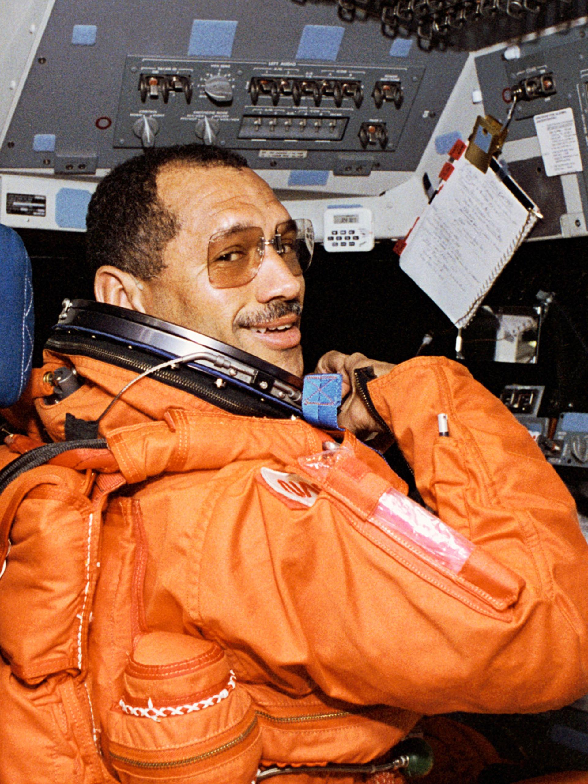 An astronaut commander (Charles F Bolden Jr) in a bright orange spacesuit sitting on the left station on the forward flight deck of the space shuttle Discovery.