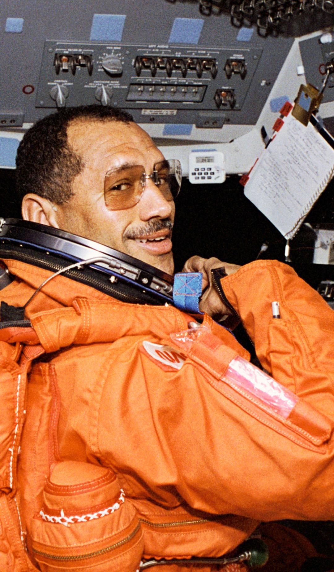 An astronaut commander (Charles F Bolden Jr) in a bright orange spacesuit sitting on the left station on the forward flight deck of the space shuttle Discovery.