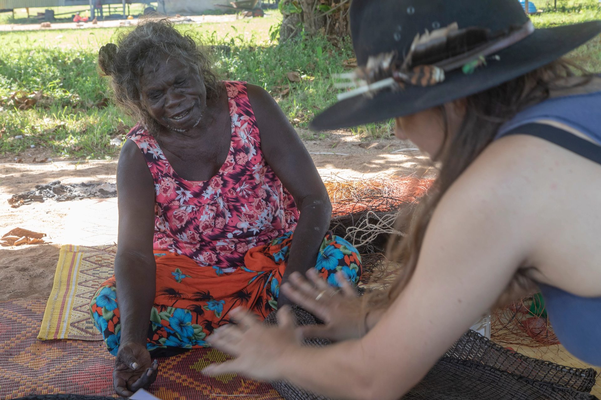 Older and younger women sit talking on woven mats