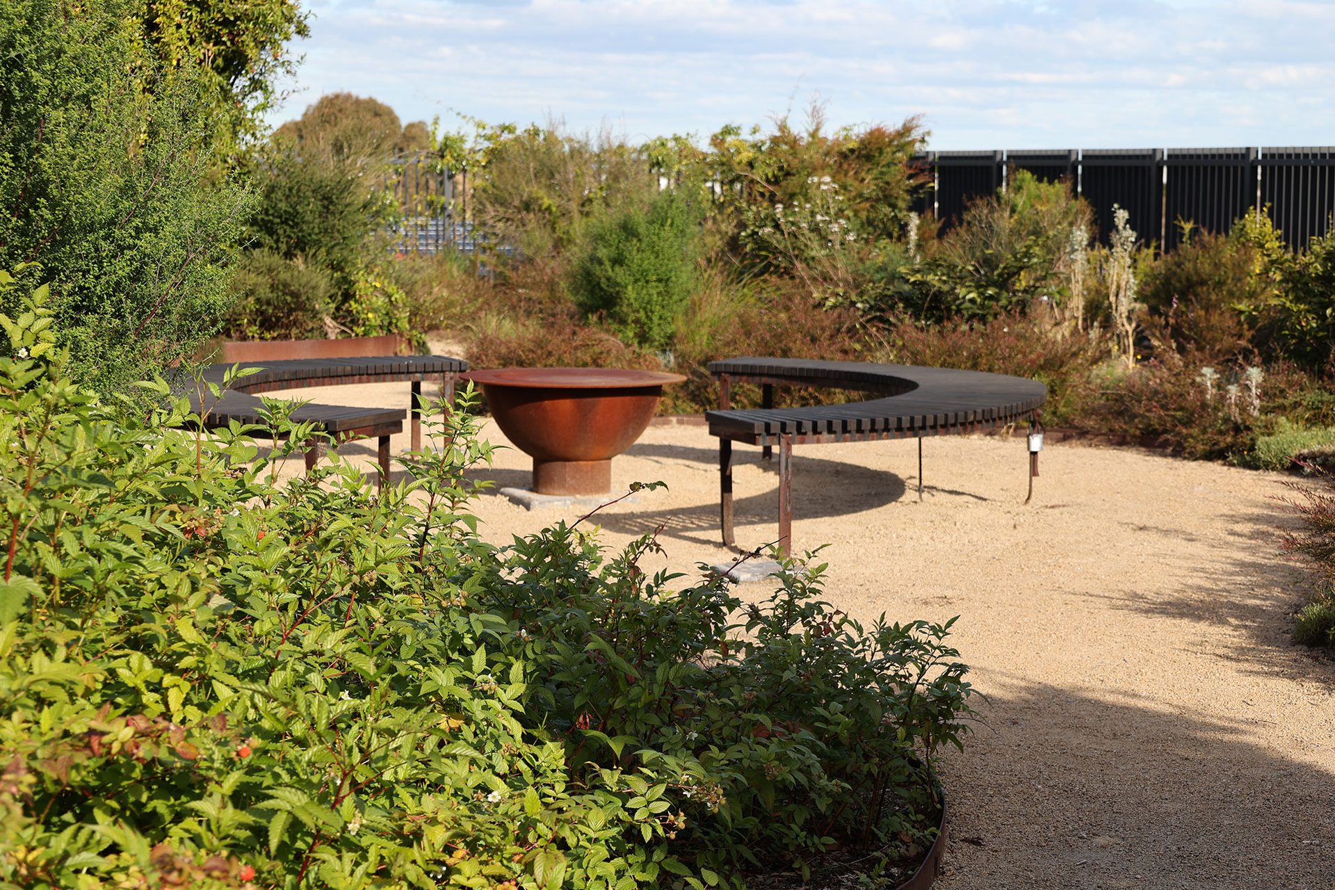 A variety of small green trees and bush surround a seating area that includes 2 hemisphere benches and a pit and sits on a gravel floor.