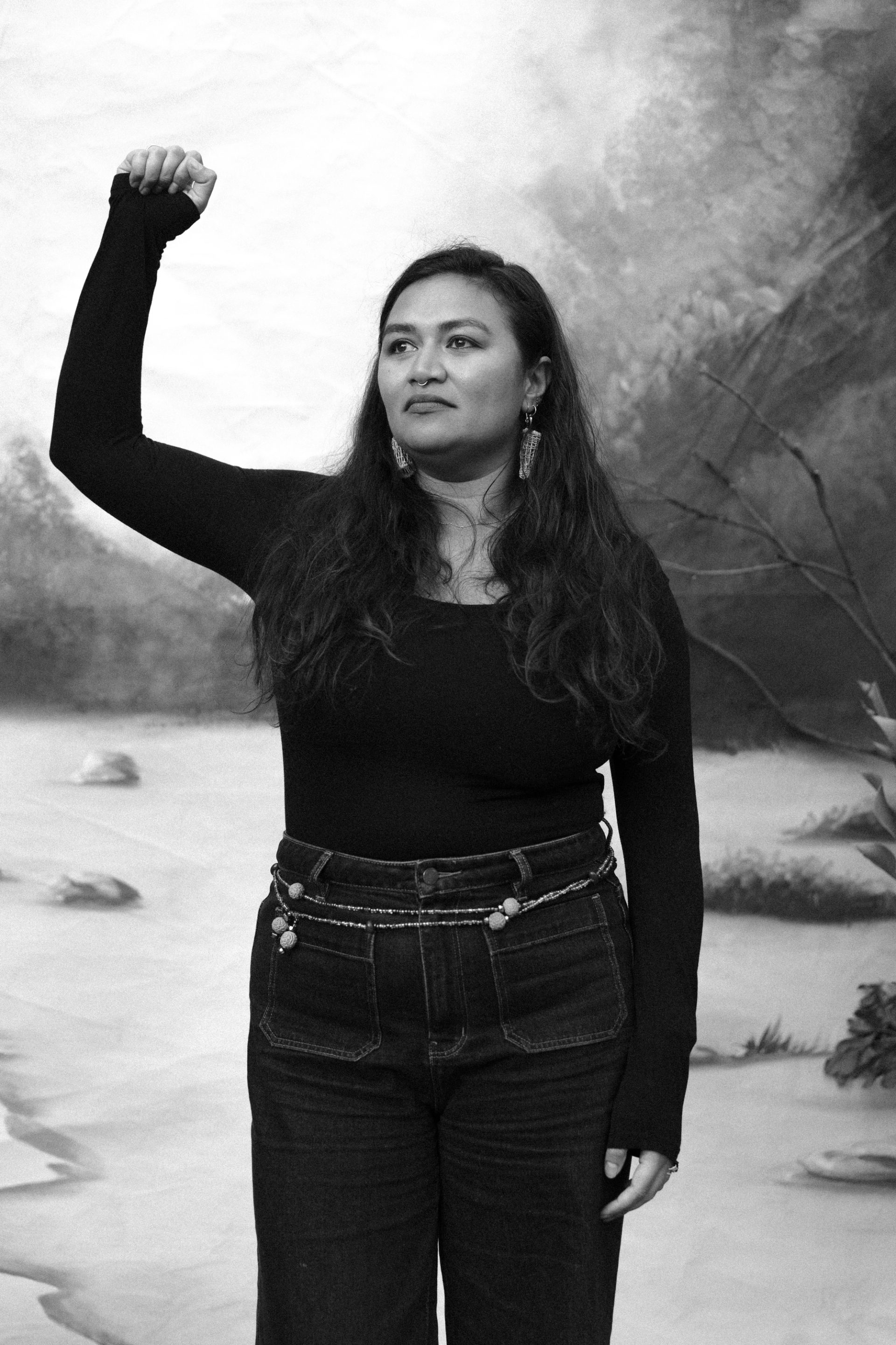 A Minyungbal woman stands in front of a painted backdrop, her right arm and fist raised.