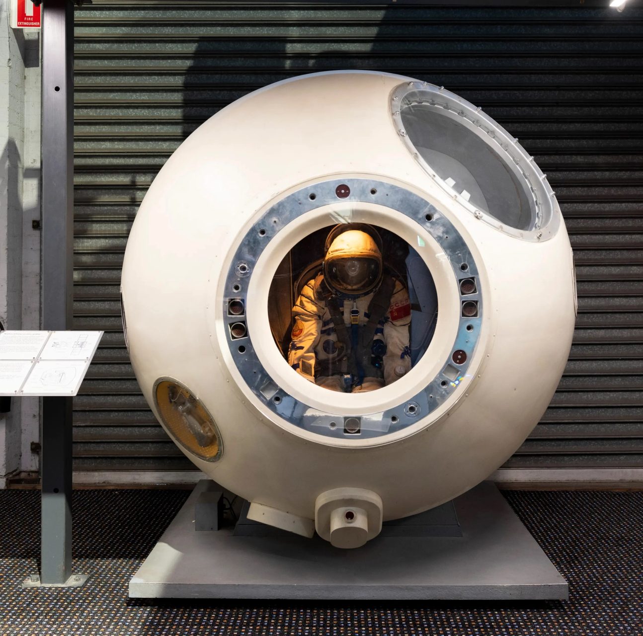 Full-scale replica of a first-generation Soyuz spacecraft re-entry module. The module is bell-shaped with two small portholes on either side of the access hatch to the 'orbital module'. The entry hatch is open showing a view into the spacecraft interior, which is reproduced in basic form, showing the layout of the main control panels and fittings. The three cosmonaut launch/landing couches are shown, two of them occupied by spacesuited mannequins representing the crew.