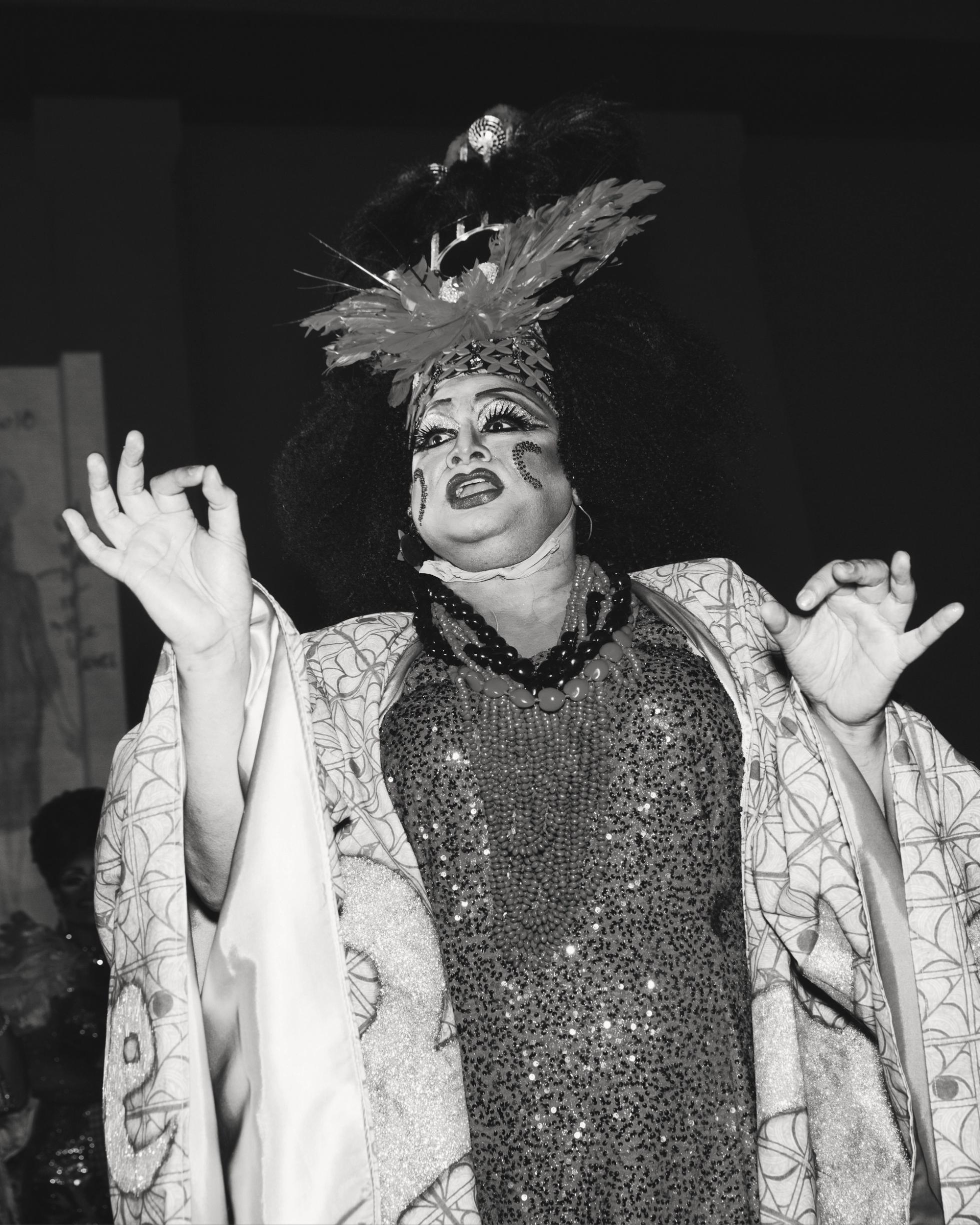 A drag queen performing wearing a patterned kimono, sequined dress and elaborate headdress.