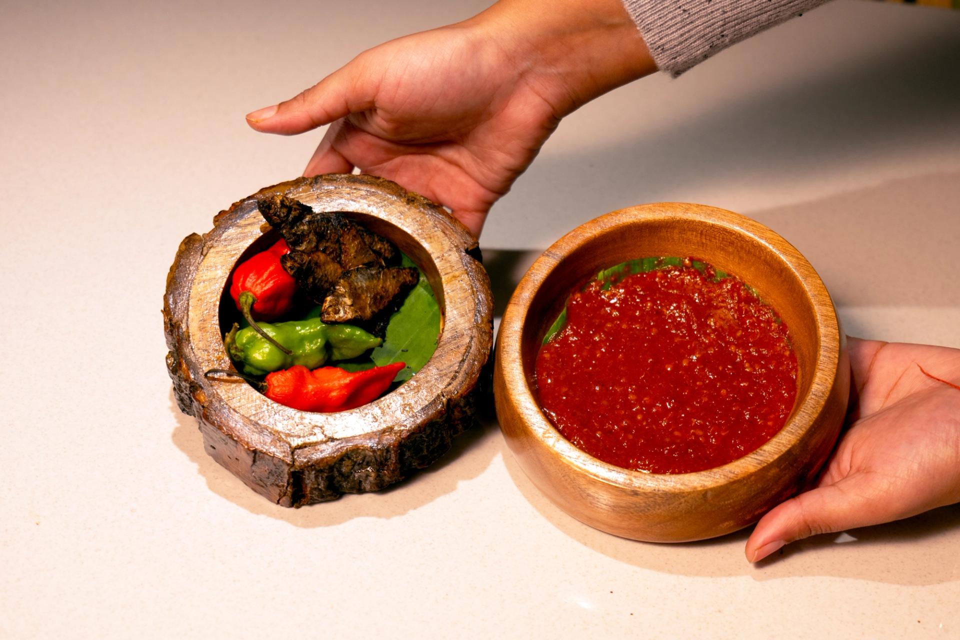 A pair of brown hands present singju ingredients in wooden bowls: fresh chilli and herbs and condiments.