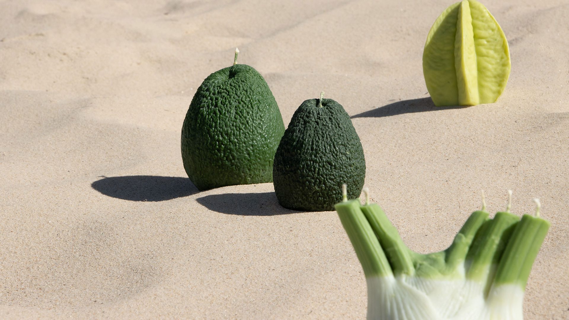 Candles resembling a starfruit, a fennel and two avocados sit on an undulating stretch of sand.