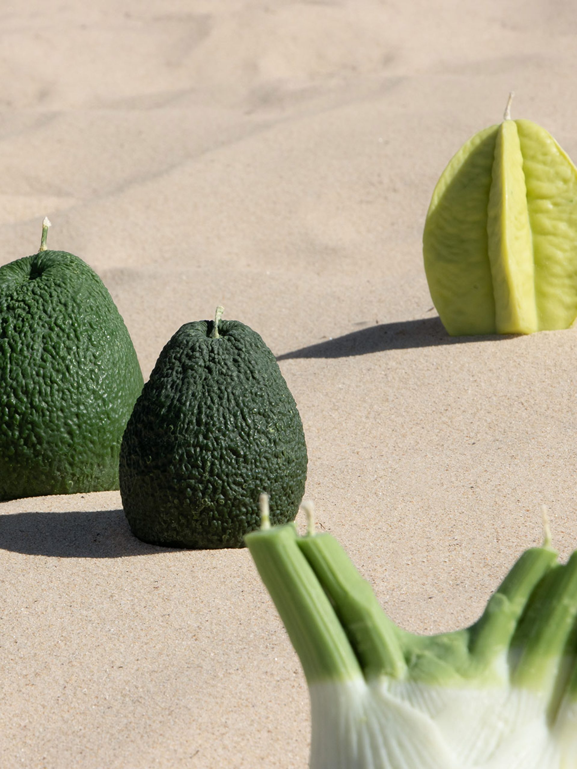 Candles resembling a starfruit, a fennel and two avocados sit on an undulating stretch of sand.
