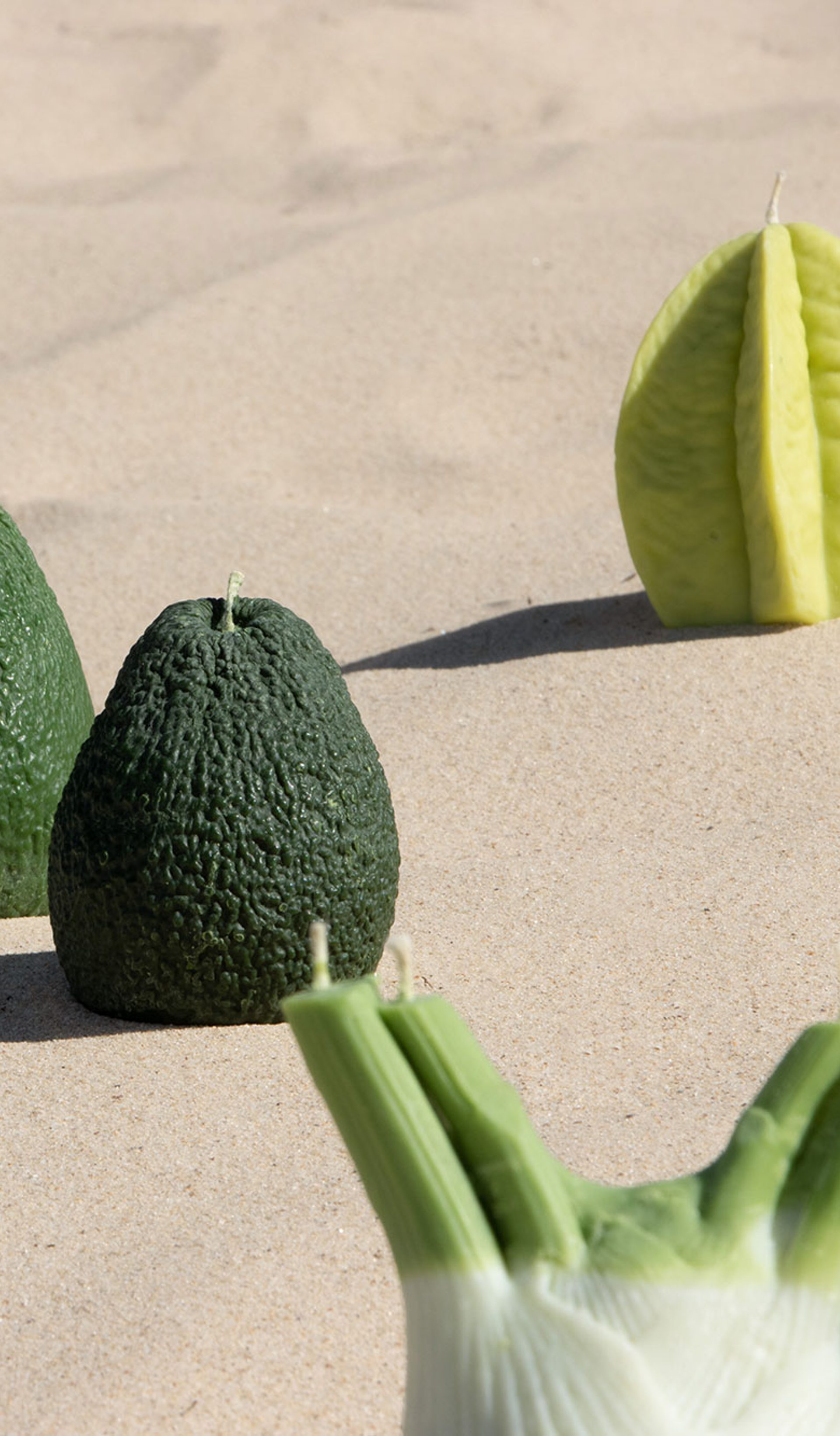 Candles resembling a starfruit, a fennel and two avocados sit on an undulating stretch of sand.