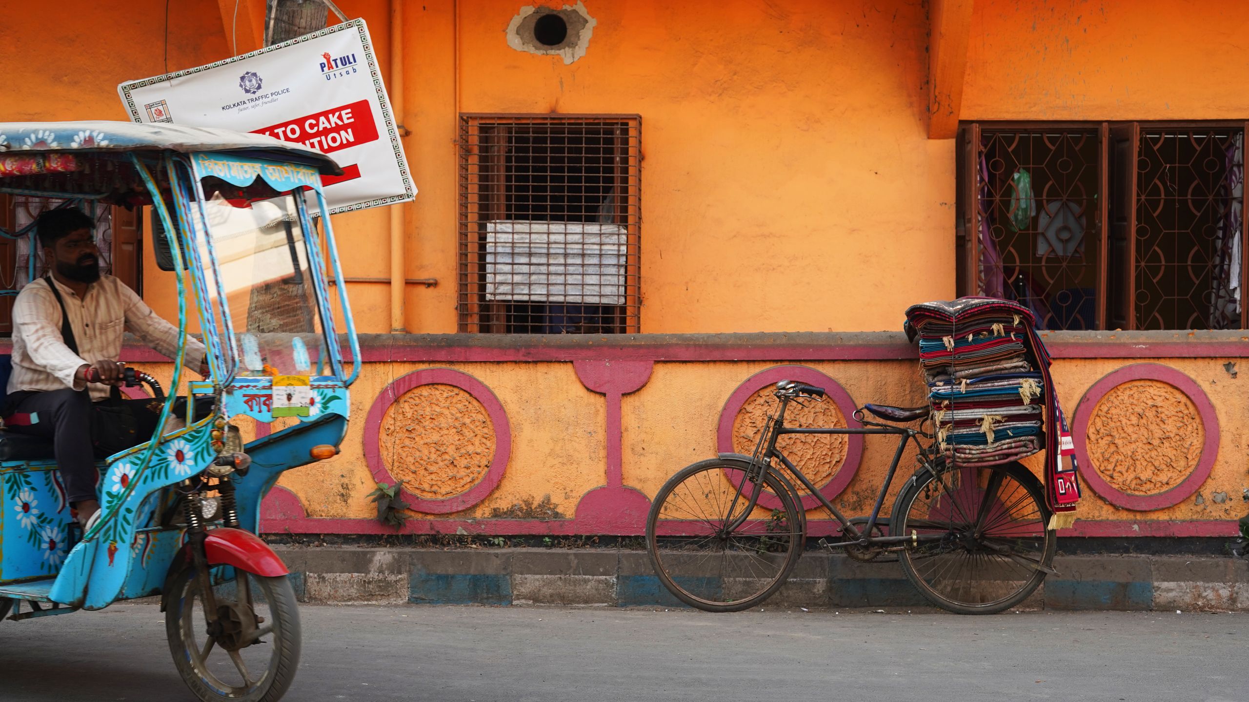 On a neighbourhood street a bicycle piled with fabrics leans against a bright orange wall. Entering the frame on the left, a man drives a blue-patterned tuk-tuk.