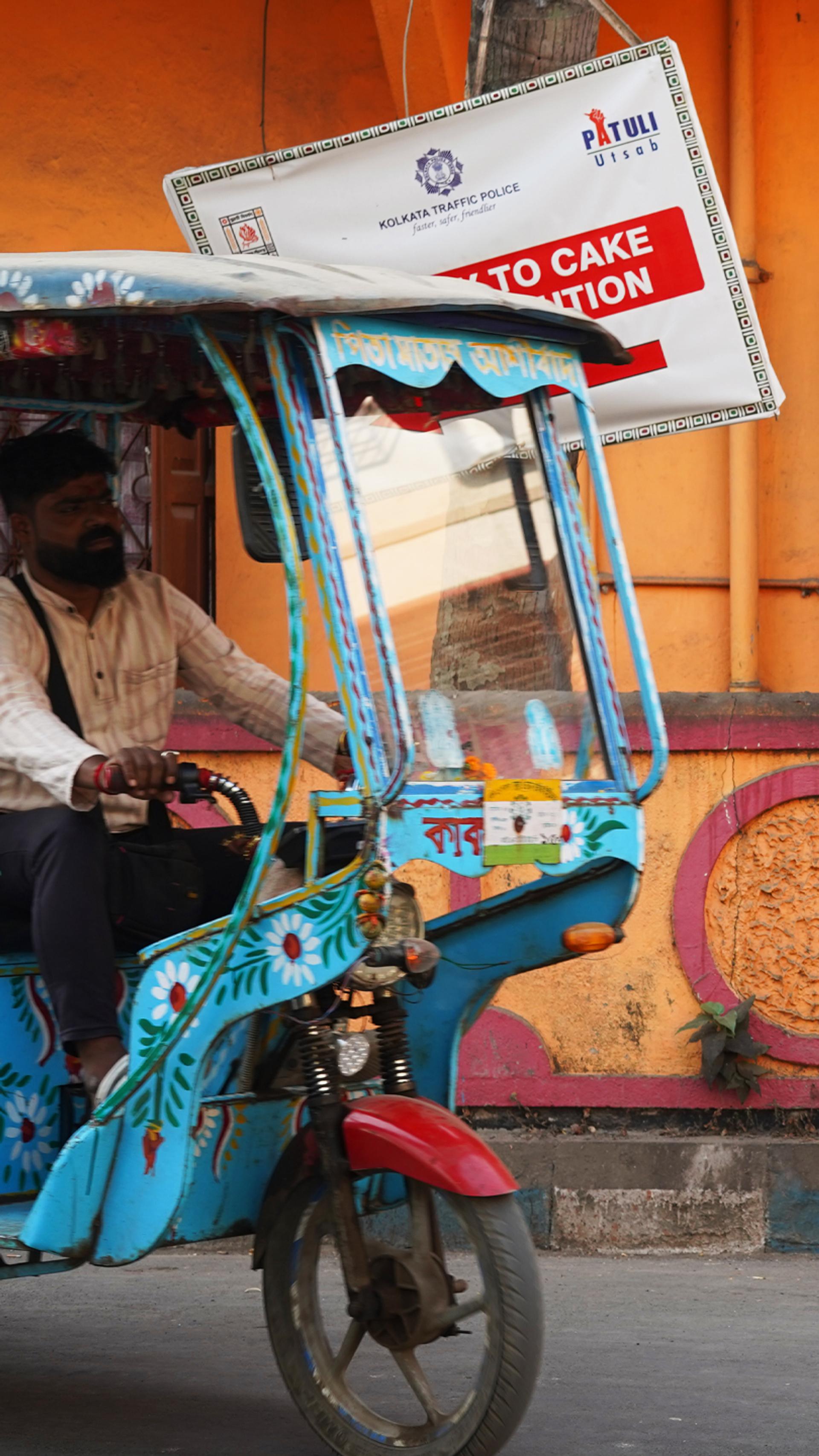 On a neighbourhood street a bicycle piled with fabrics leans against a bright orange wall. Entering the frame on the left, a man drives a blue-patterned tuk-tuk.