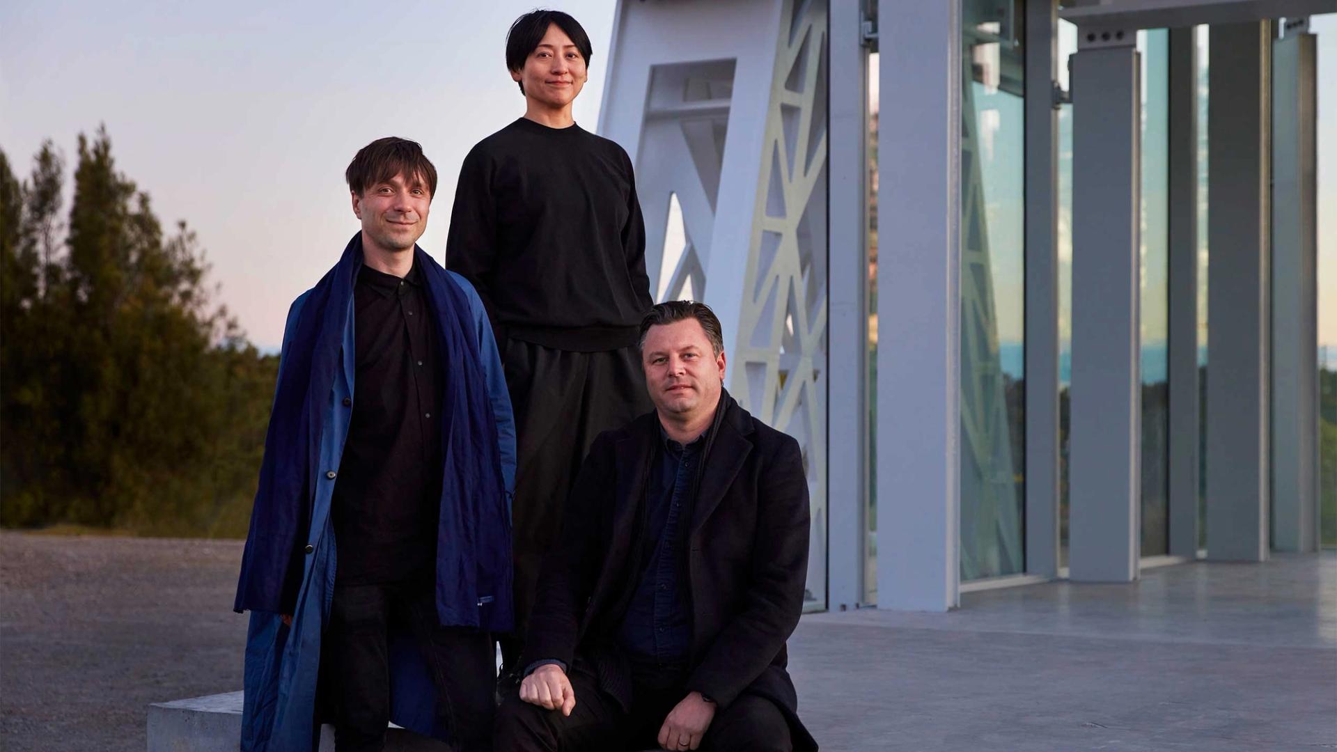 Three people (Powerhouse Parramatta architects Nicolas Moreau, Hiroko Kusunoki and Steve Toia) posed in front of a large white beam structure looking towards the camera.