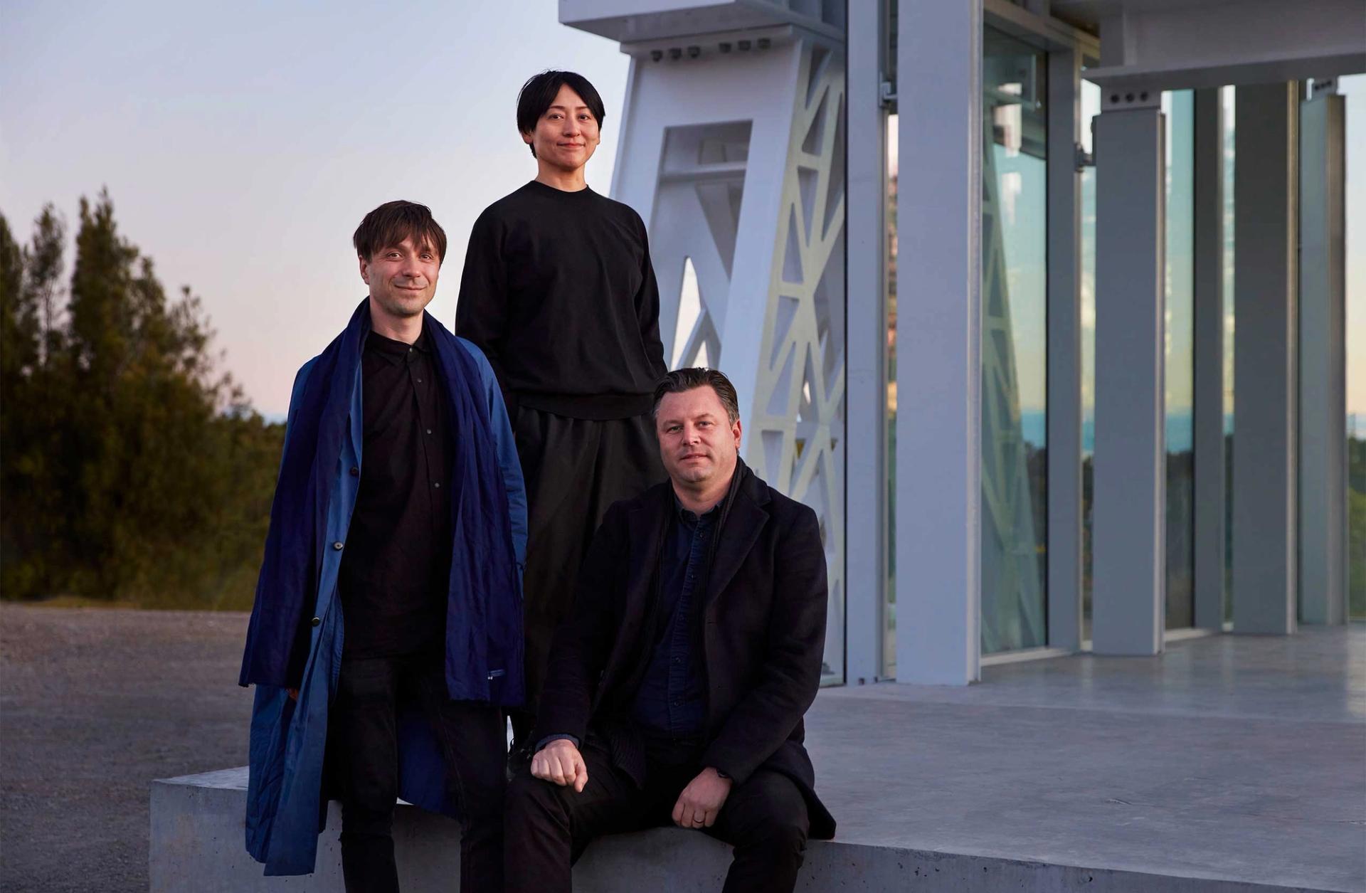 Three people (Powerhouse Parramatta architects Nicolas Moreau, Hiroko Kusunoki and Steve Toia) posed in front of a large white beam structure looking towards the camera.