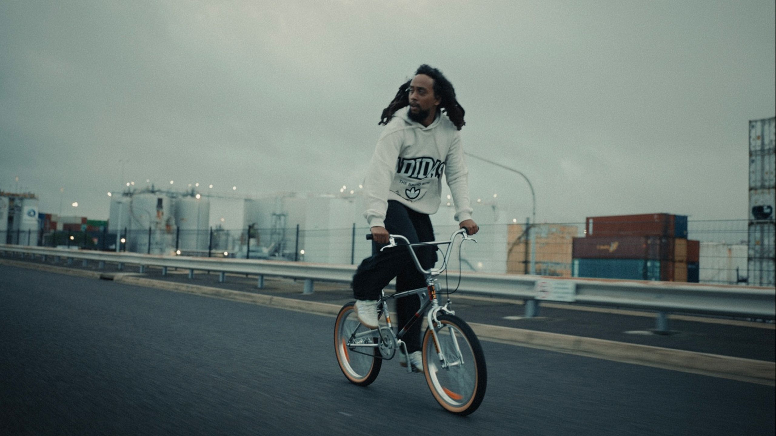 A photograph of a young person on a bicycle, riding on a highway, their hair blowing in the wind. They wear a white hoodie.