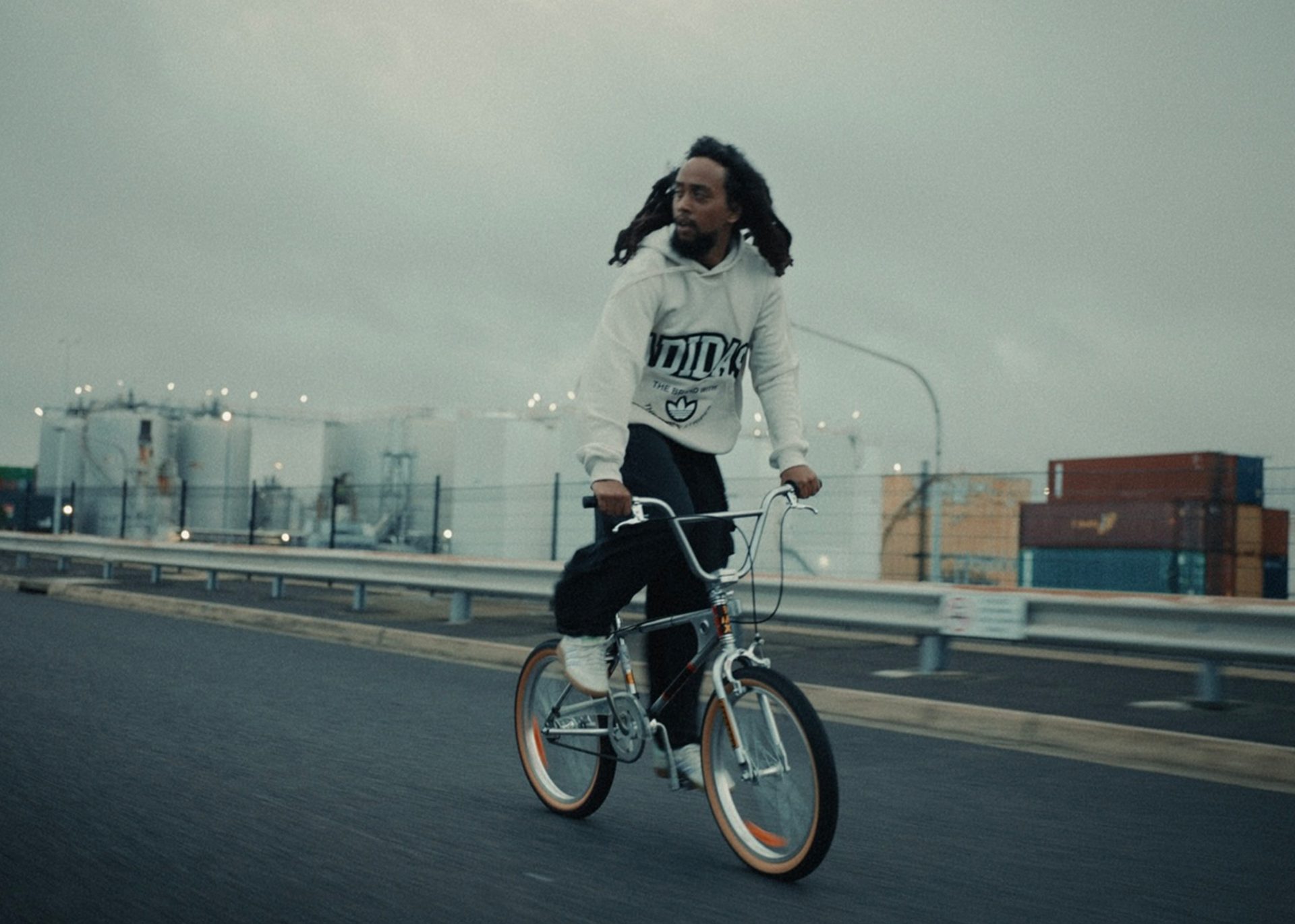 A photograph of a young person on a bicycle, riding on a highway, their hair blowing in the wind. They wear a white hoodie.