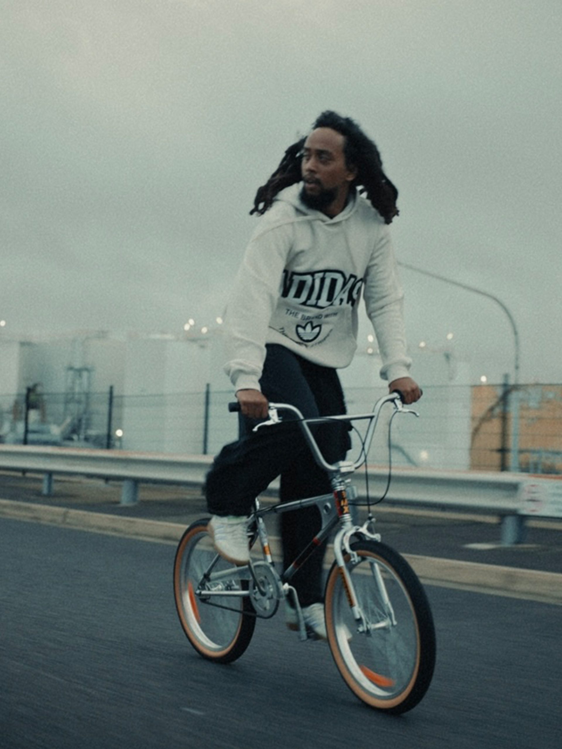 A photograph of a young person on a bicycle, riding on a highway, their hair blowing in the wind. They wear a white hoodie.