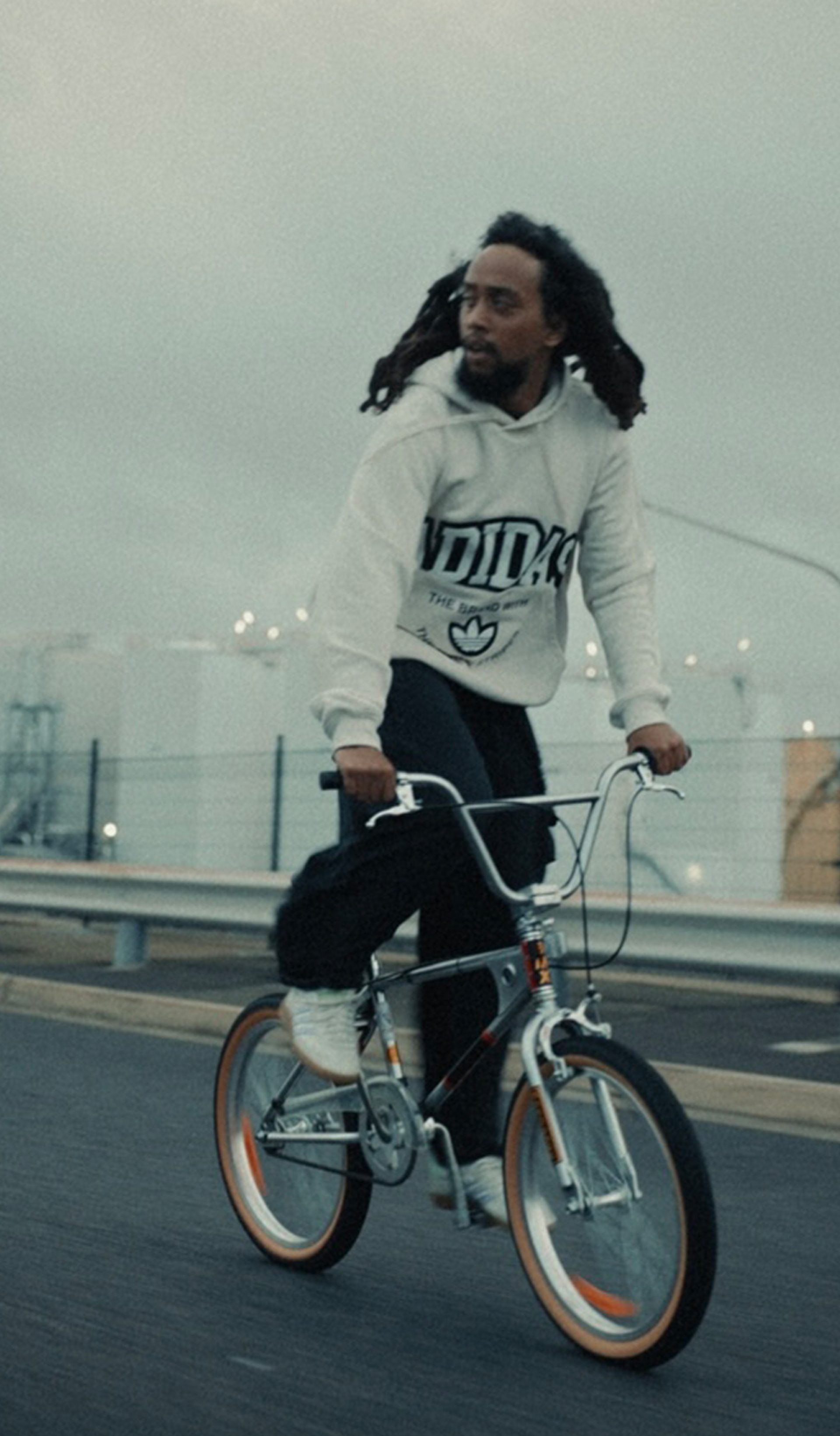 A photograph of a young person on a bicycle, riding on a highway, their hair blowing in the wind. They wear a white hoodie.