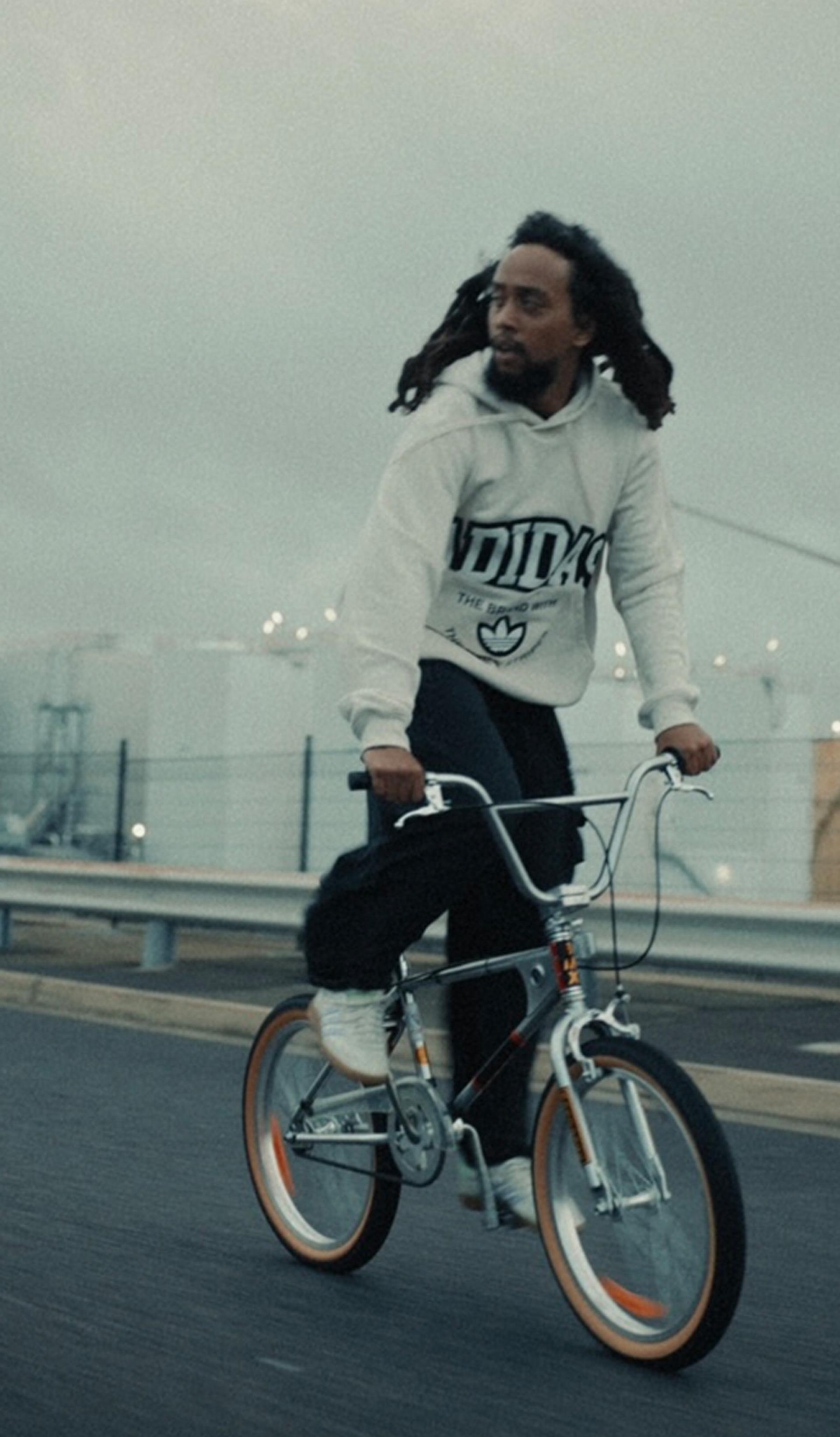 A photograph of a young person on a bicycle, riding on a highway, their hair blowing in the wind. They wear a white hoodie.