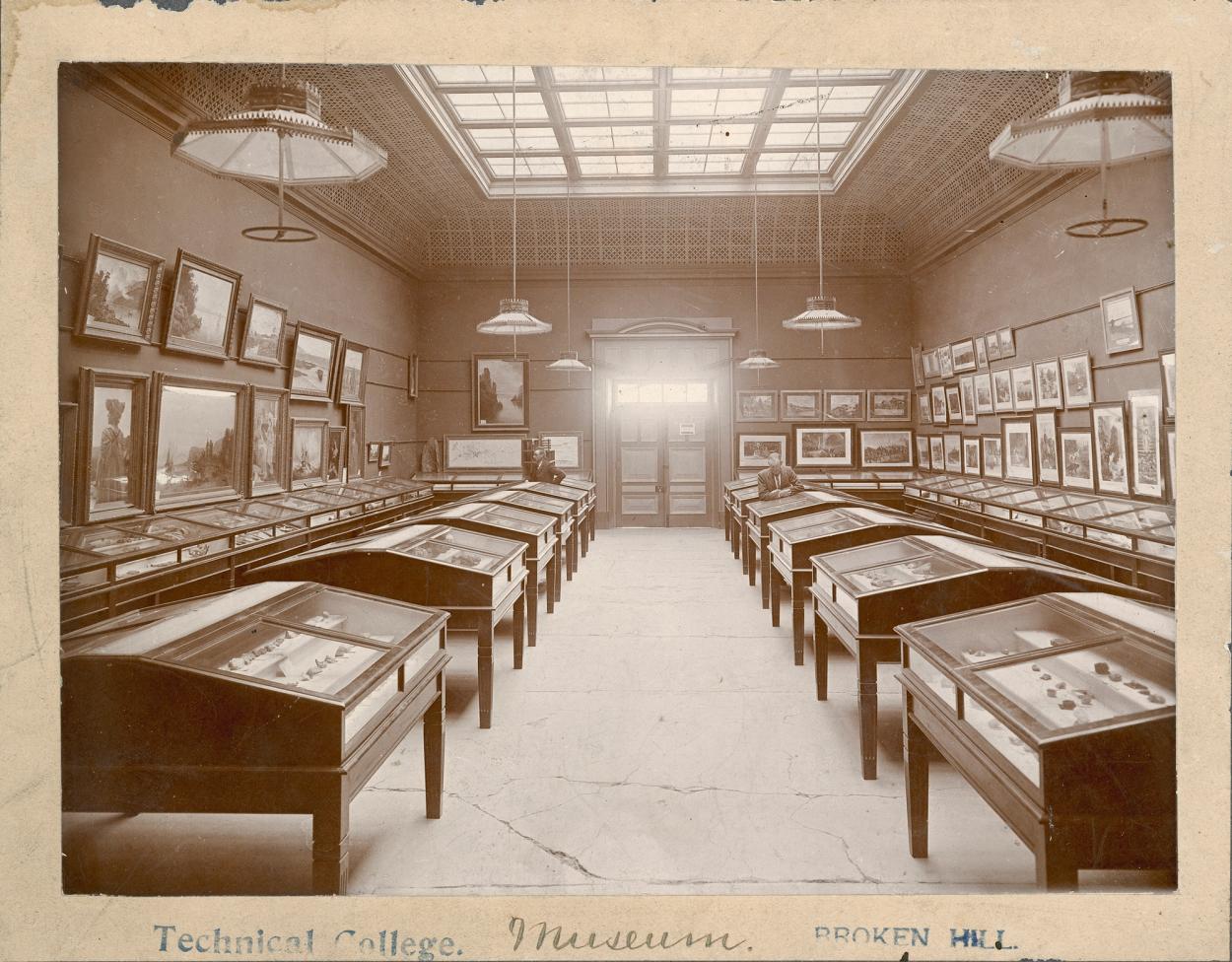 Sepia photograph of a museum display with cabinets and pictures hanging from either wall