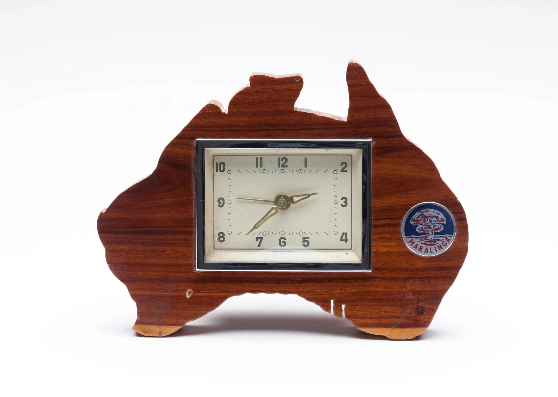 A mechanical clock set in a solid block casing of varnished mulga wood, in the shape of the Australian mainland. The inset rectangular clock face is cream with a border of silver metal. The words 'West Germany' appear at the bottom of the face. To the right of the clock face an enameled metal disc is attached to the wood. It has a silver background with a blue and red illustration of an atomic explosion with a double mushroom cloud above the word 'Maralinga' in red.