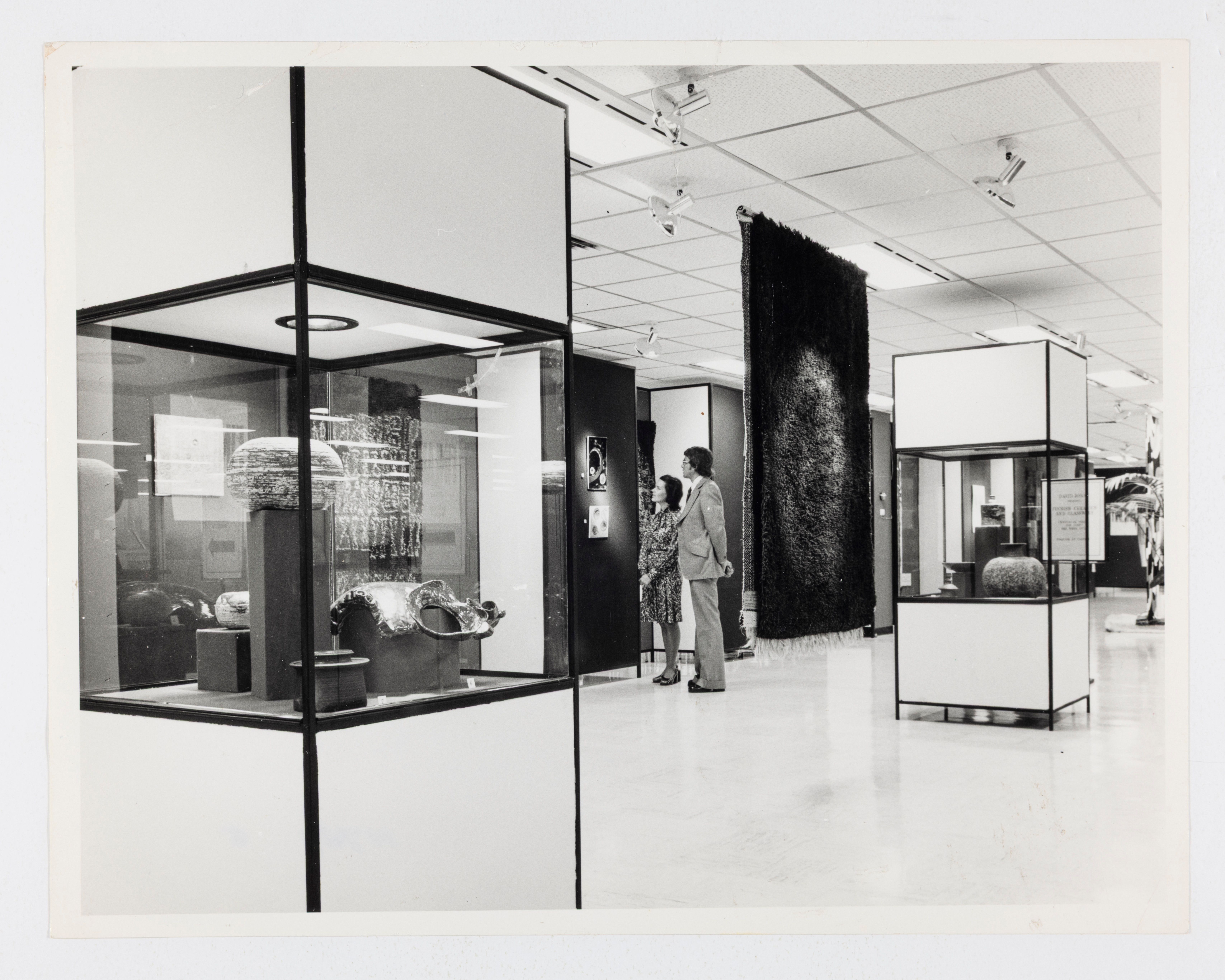 A department store interior featuring objects displayed in large glass cases and a hanging textile. At the centre back, a man and a woman view a small artwork, mounted on the wall.
