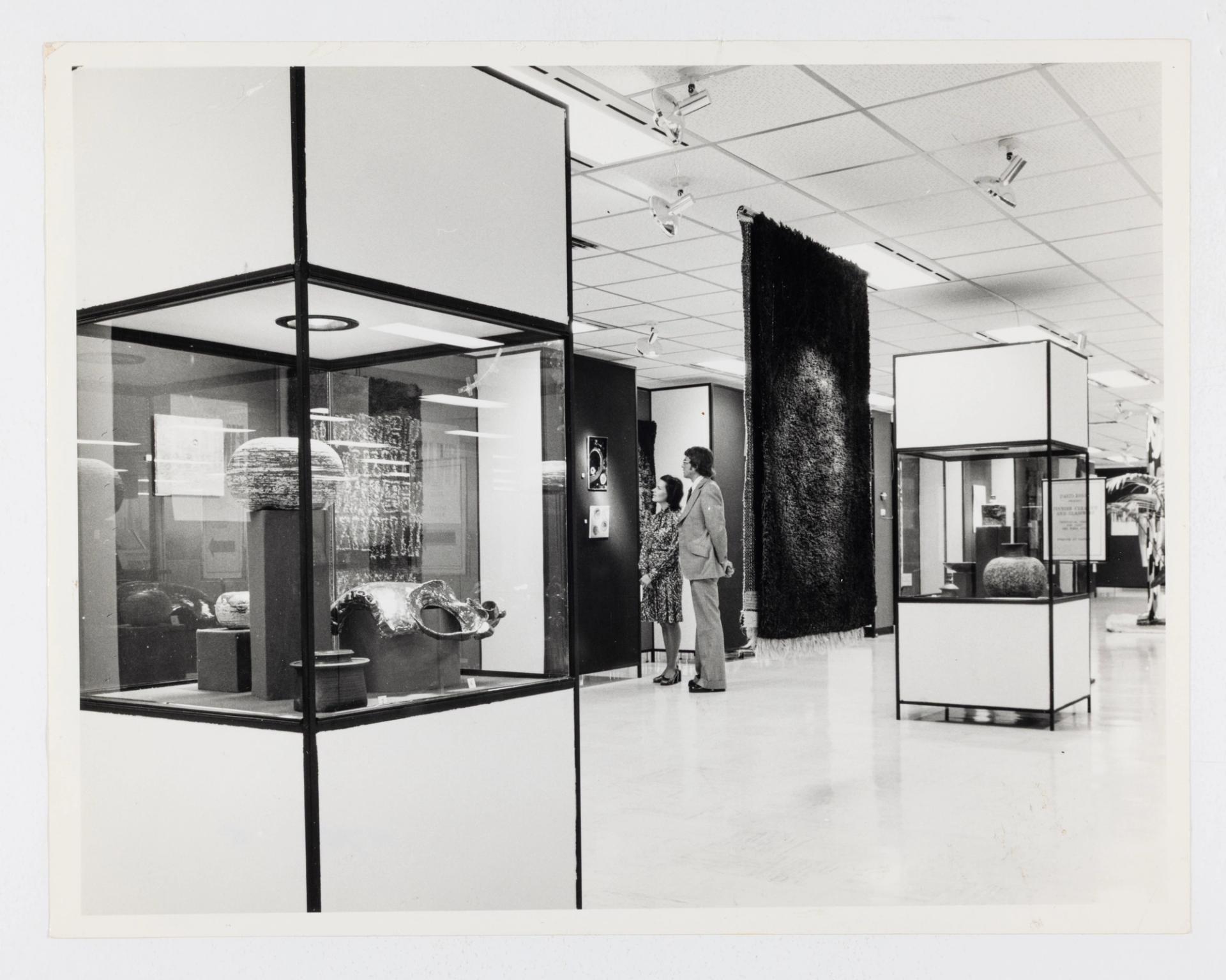 A department store interior featuring objects displayed in large glass cases and a hanging textile. At the centre back, a man and a woman view a small artwork, mounted on the wall.