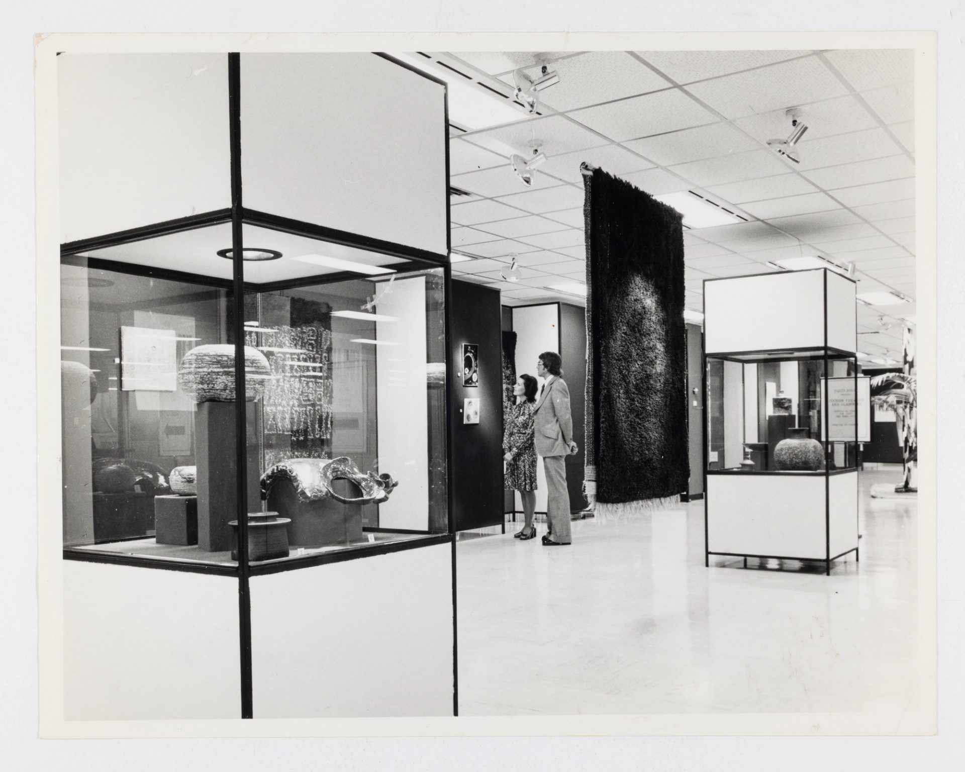 A department store interior featuring objects displayed in large glass cases and a hanging textile. At the centre back, a man and a woman view a small artwork, mounted on the wall.
