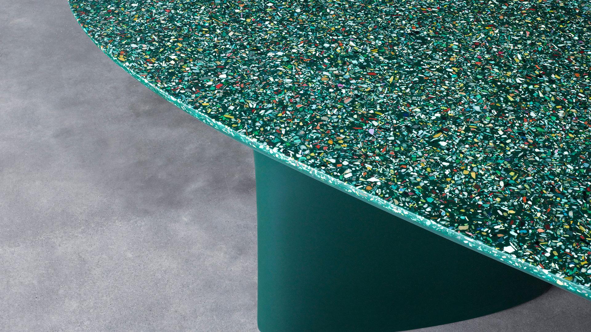 Green table on a concrete floor. The top of the table is an irregularly shaped ellipsis, showing the terrazzo, ocean plastic composition of the table, with flecks of colour amid the overall green appearance.