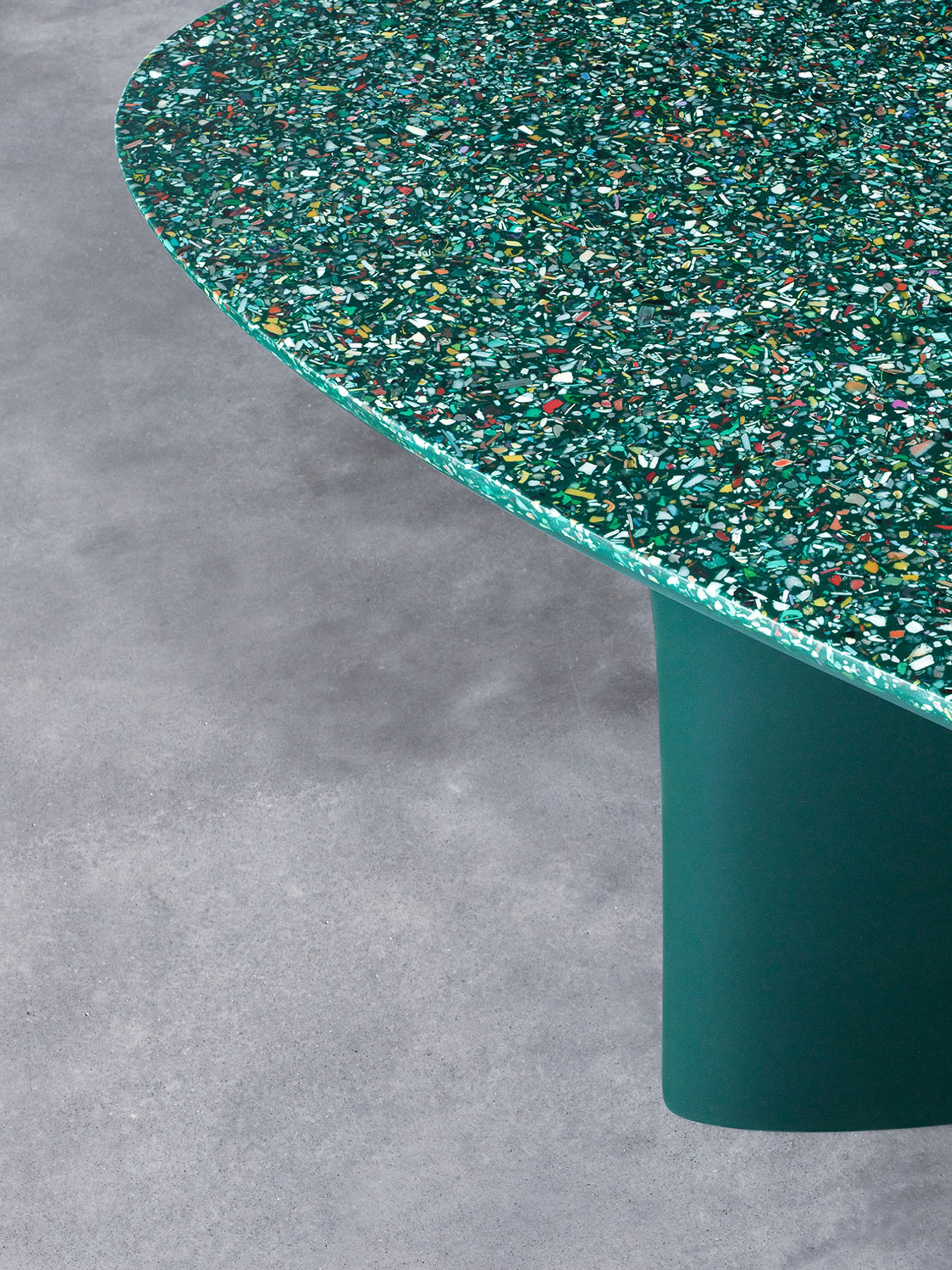 Green table on a concrete floor. The top of the table is an irregularly shaped ellipsis, showing the terrazzo, ocean plastic composition of the table, with flecks of colour amid the overall green appearance.