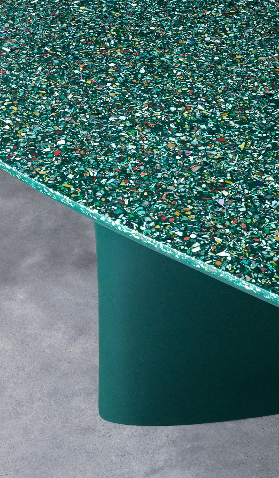 Green table on a concrete floor. The top of the table is an irregularly shaped ellipsis, showing the terrazzo, ocean plastic composition of the table, with flecks of colour amid the overall green appearance.
