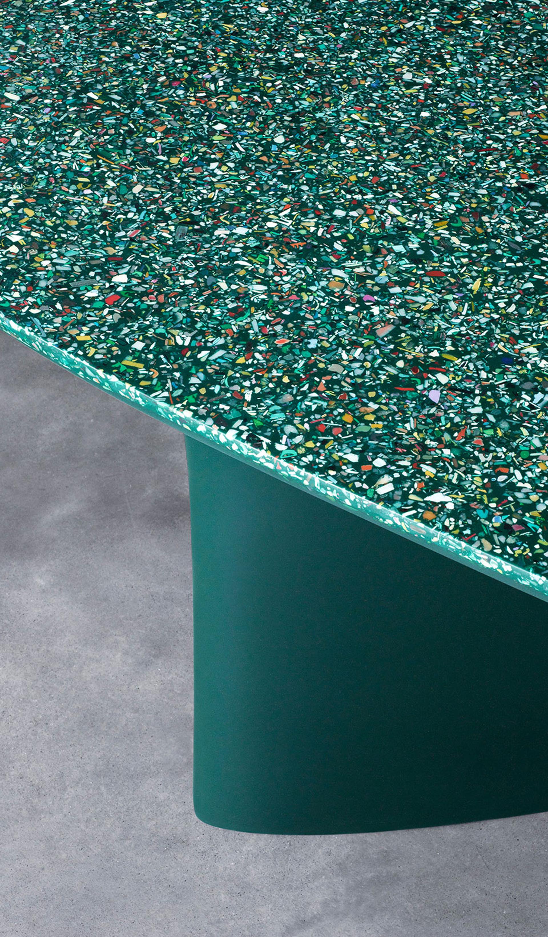 Green table on a concrete floor. The top of the table is an irregularly shaped ellipsis, showing the terrazzo, ocean plastic composition of the table, with flecks of colour amid the overall green appearance.