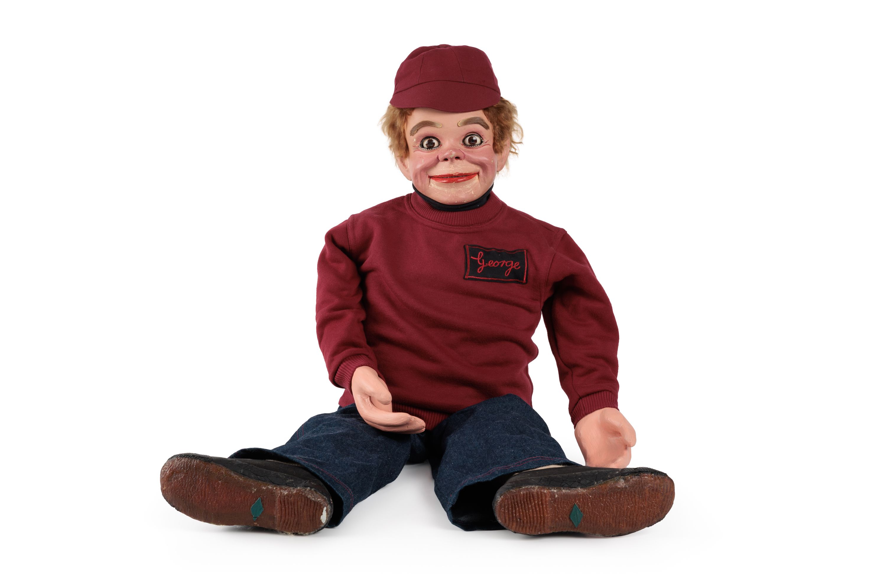 Ventriloquist figure with blonde or light brown coloured hair and eyebrows, brown eyes, red lips, white teeth and rosy cheeks.