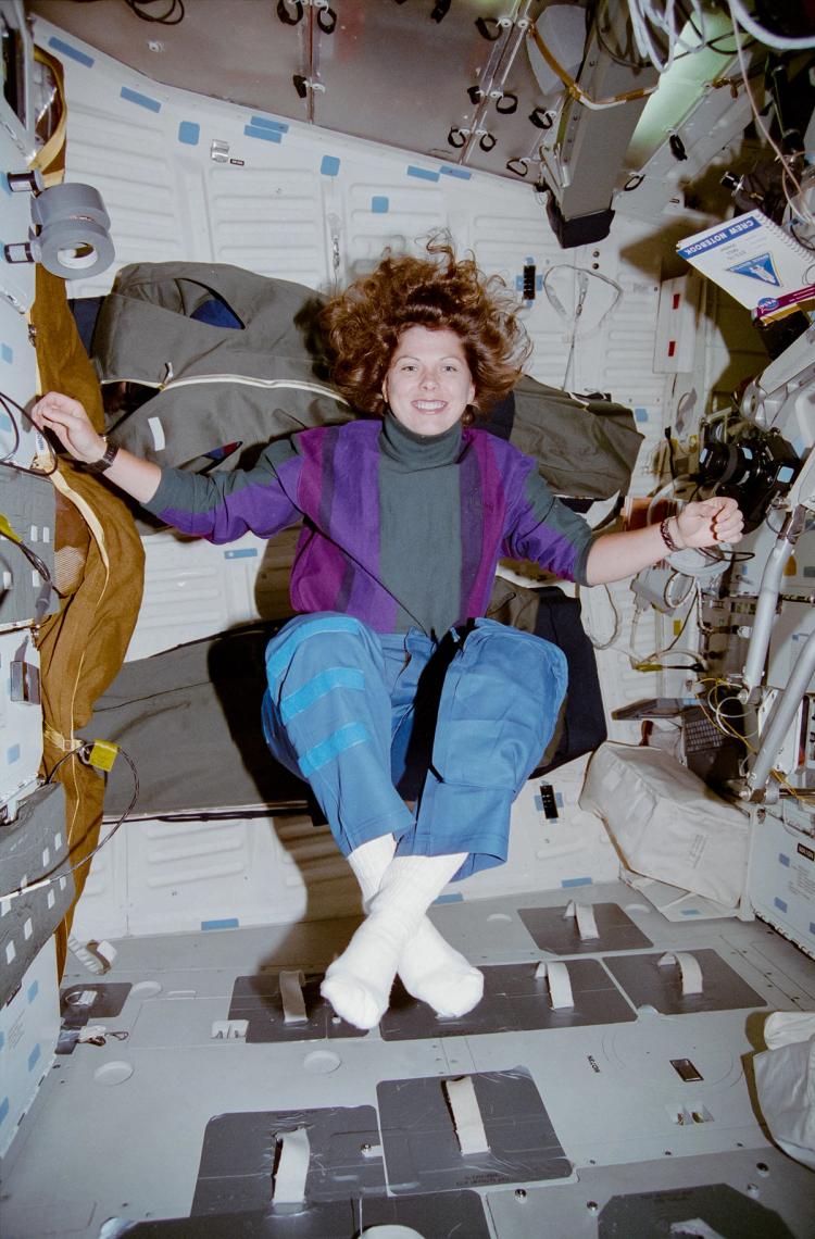 Woman wearing purple jumper, blue pants and white socks floats in mid air in a space capsule in a yoga position