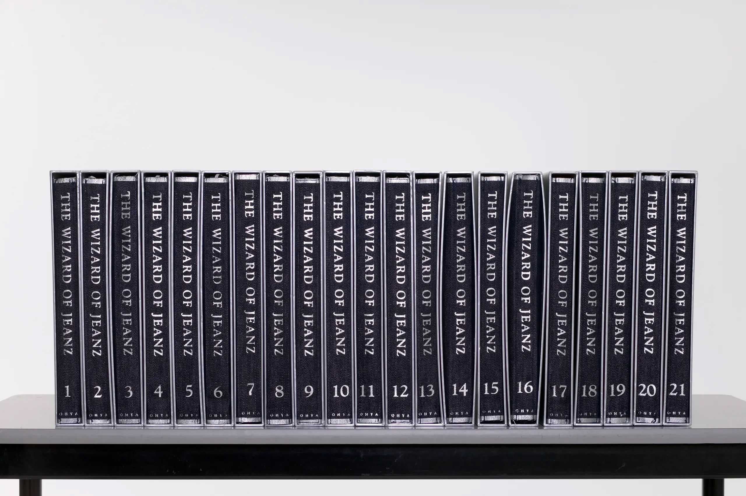 The 'Wizard of Jeanz' collection is a series of twenty-one fabric books that fold out to become different kinds of women's garments. The clothes are sewn into the inside 'spine' of a denim-look fabric booklet which can be folded to fit inside a silver cardboard slip cover. The slipcovers can be placed together and shelved as books. The text 'The Wizard of Jeanz', the volume number of each book and the designer's surname Ohya is printed along the exterior spine of the folded material in silver. The text 'The Wizard of Jeanz' and the surname Ohya is also embossed along the spine of the slip cover. As the series of garments progresses, the clothes change gradually from denim-style clothing to floral cotton.
